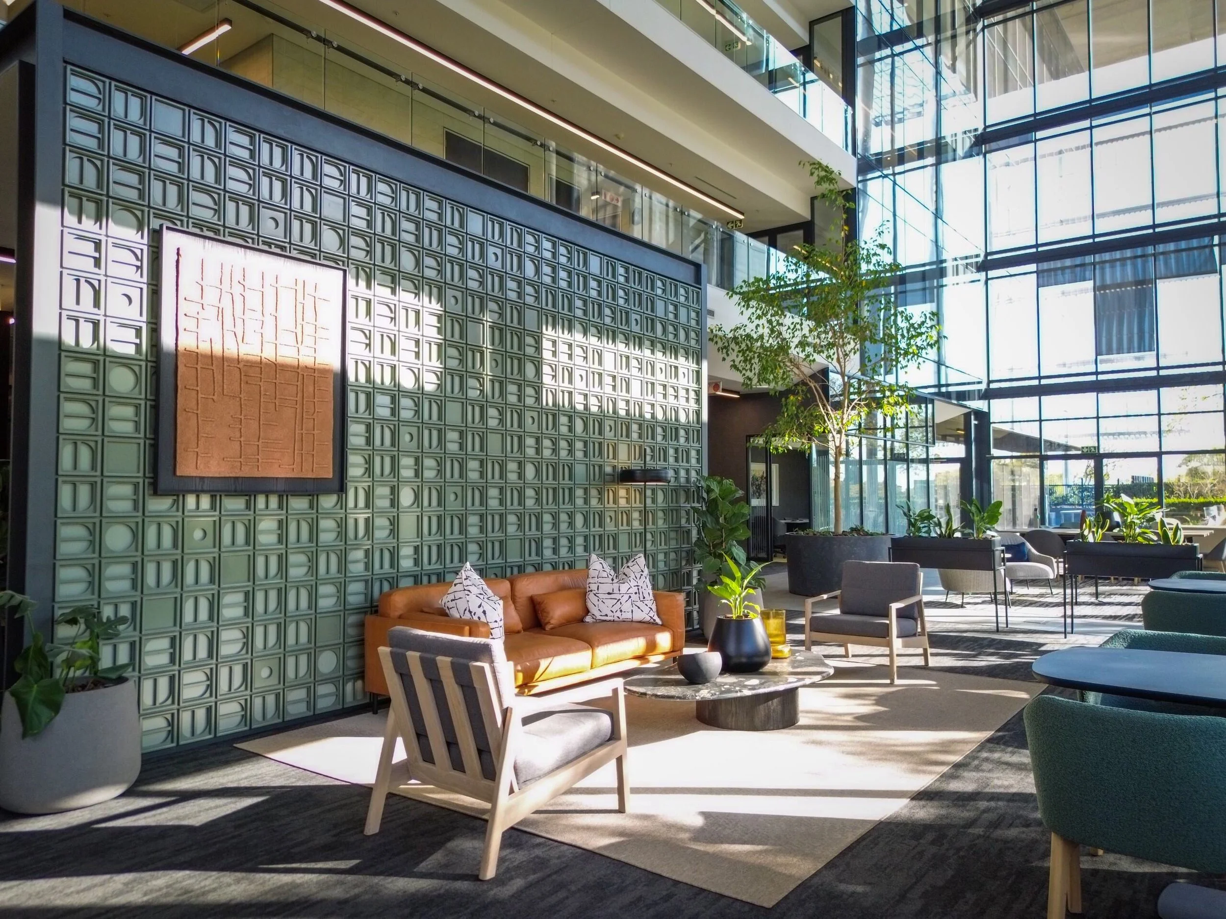 Modern hotel lobby with large glass windows, green plants, and stylish furniture, including a tan leather sofa and gray chairs, illuminated by natural sunlight.