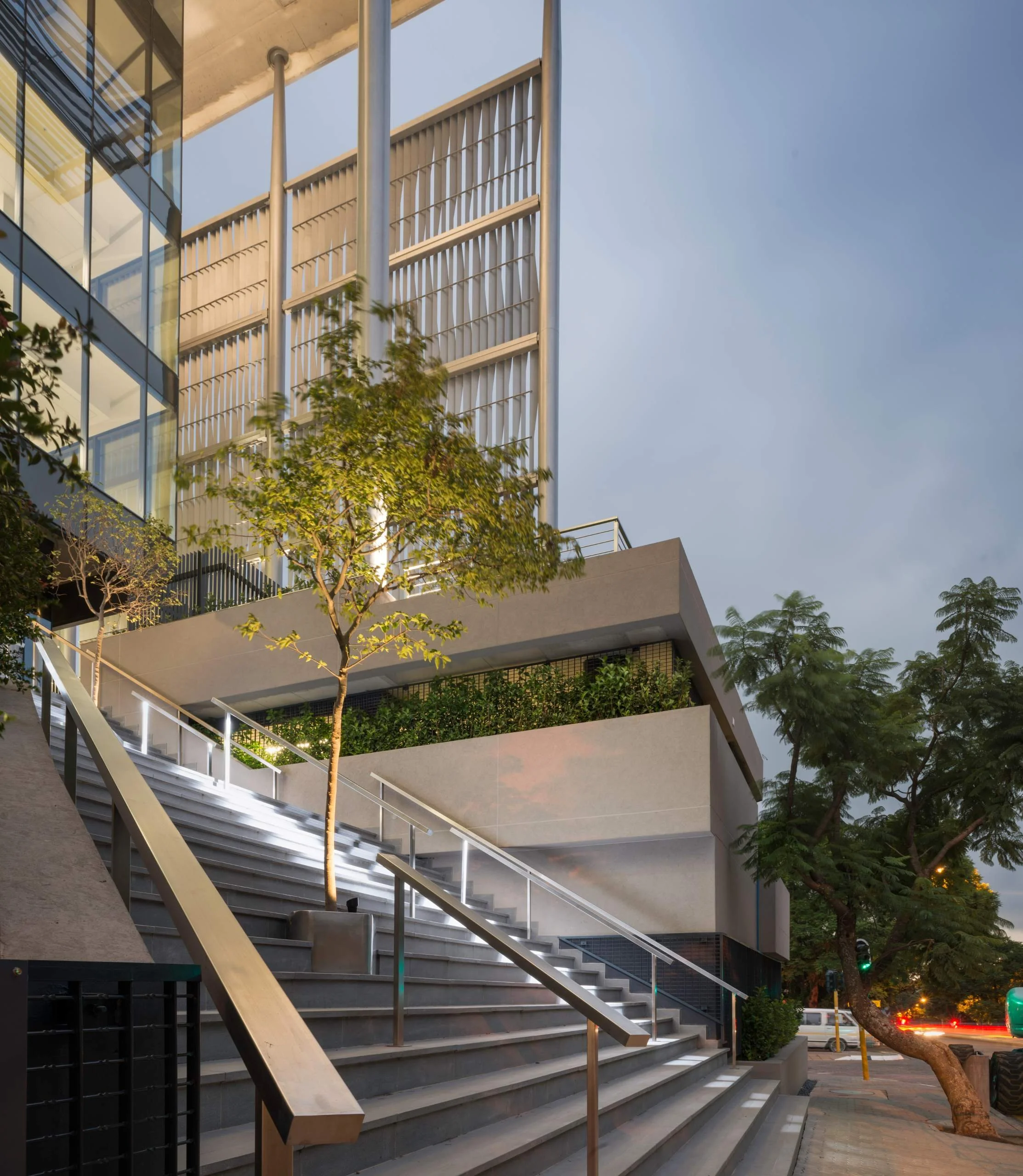 Modern multi-story building with glass windows and a staircase leading up to the entrance, surrounded by trees on a city street at dusk.