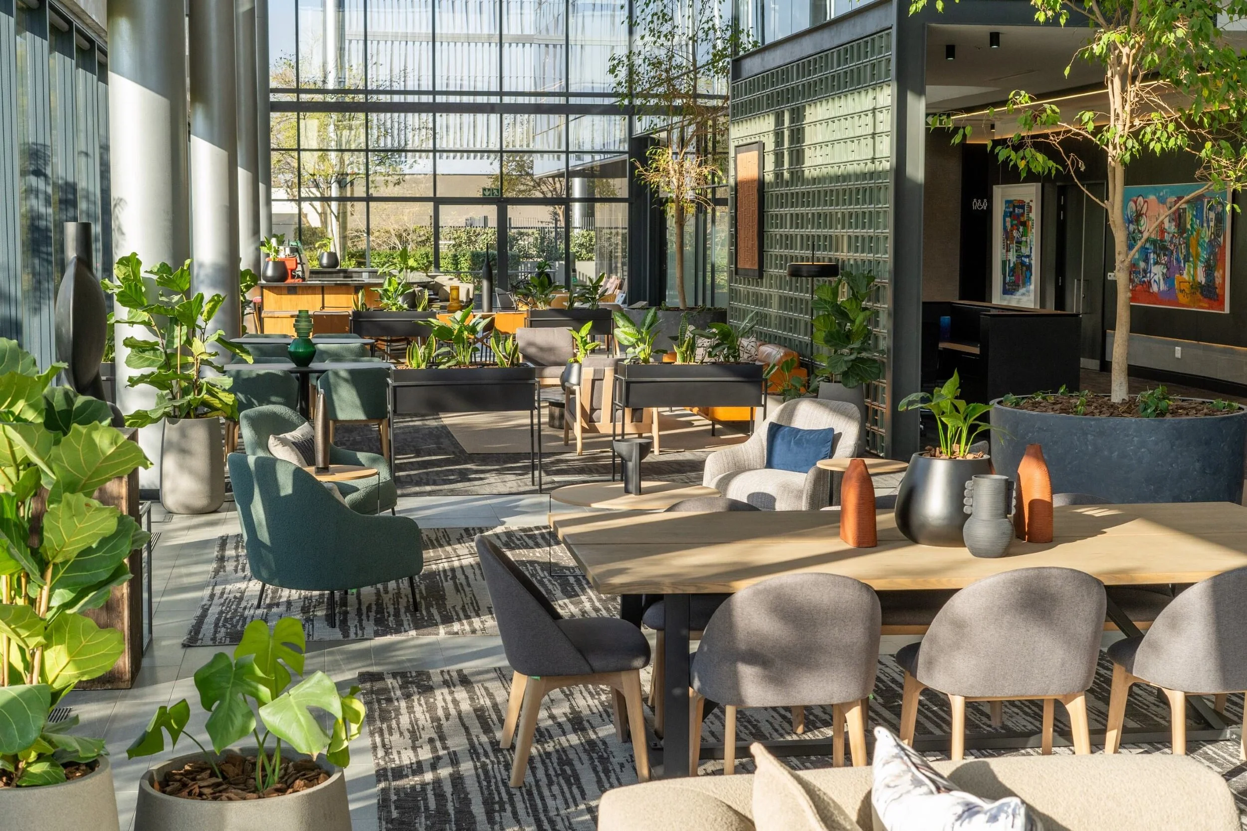 Bright, modern indoor lounge area with large glass windows, various seating options including armchairs and sofas, numerous potted plants, and contemporary decor.