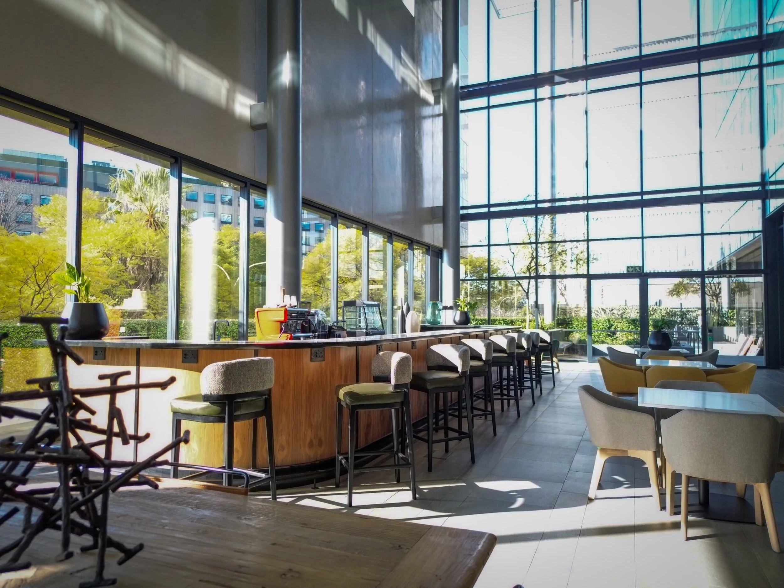 Bright, modern interior space with tall glass windows, a curved wooden bar with barstools, and seating area with beige and yellow chairs.