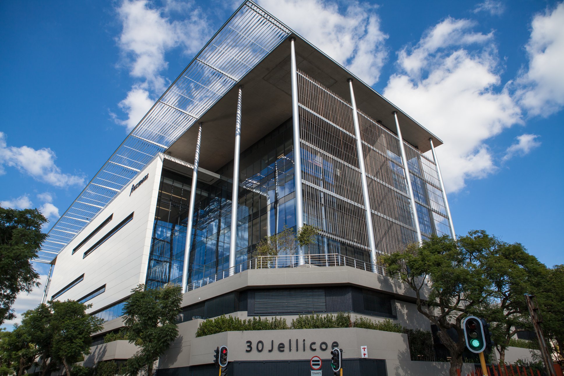Modern multi-story office building with glass facade and metal accents, labeled 30 Jellicoe, with blue sky and scattered clouds overhead, trees in front, and traffic lights at street level.