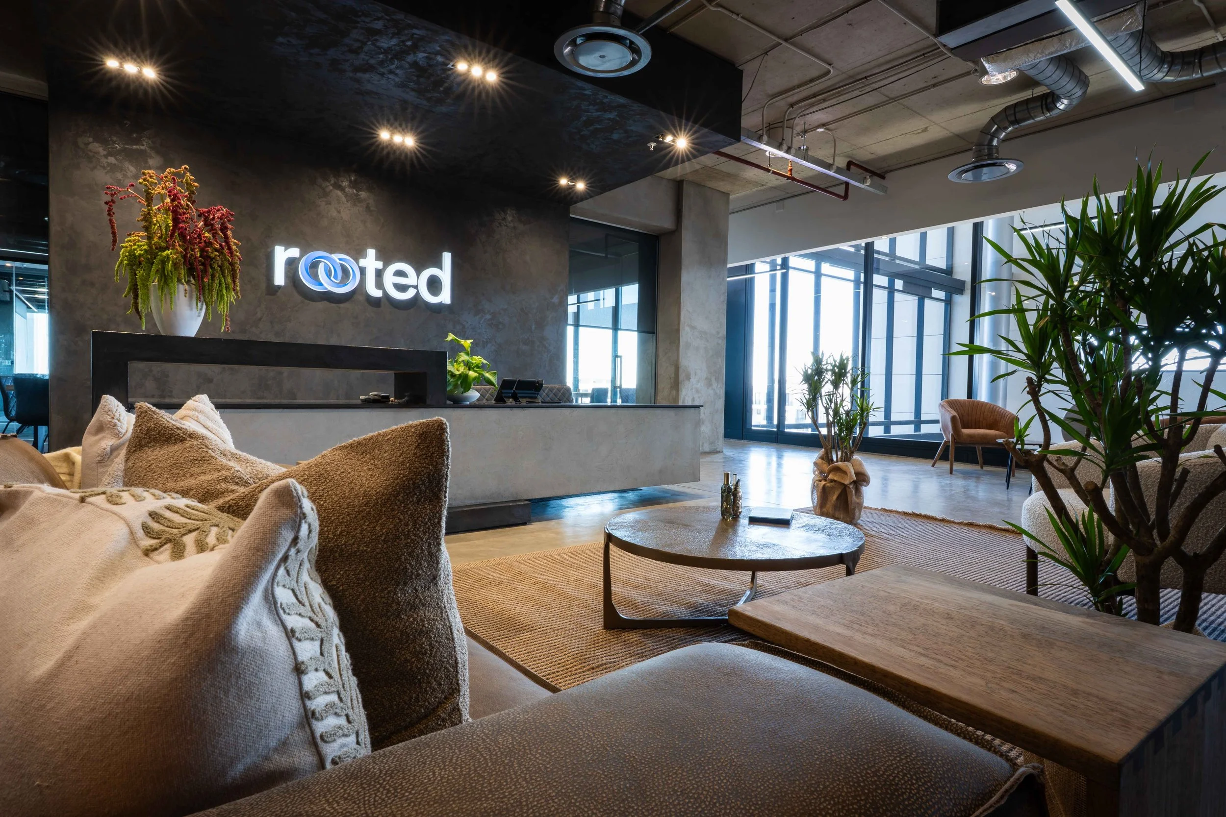 Modern hotel lobby with beige and brown furniture, plants, a reception desk, and a sign that says 'rooted' on the wall.