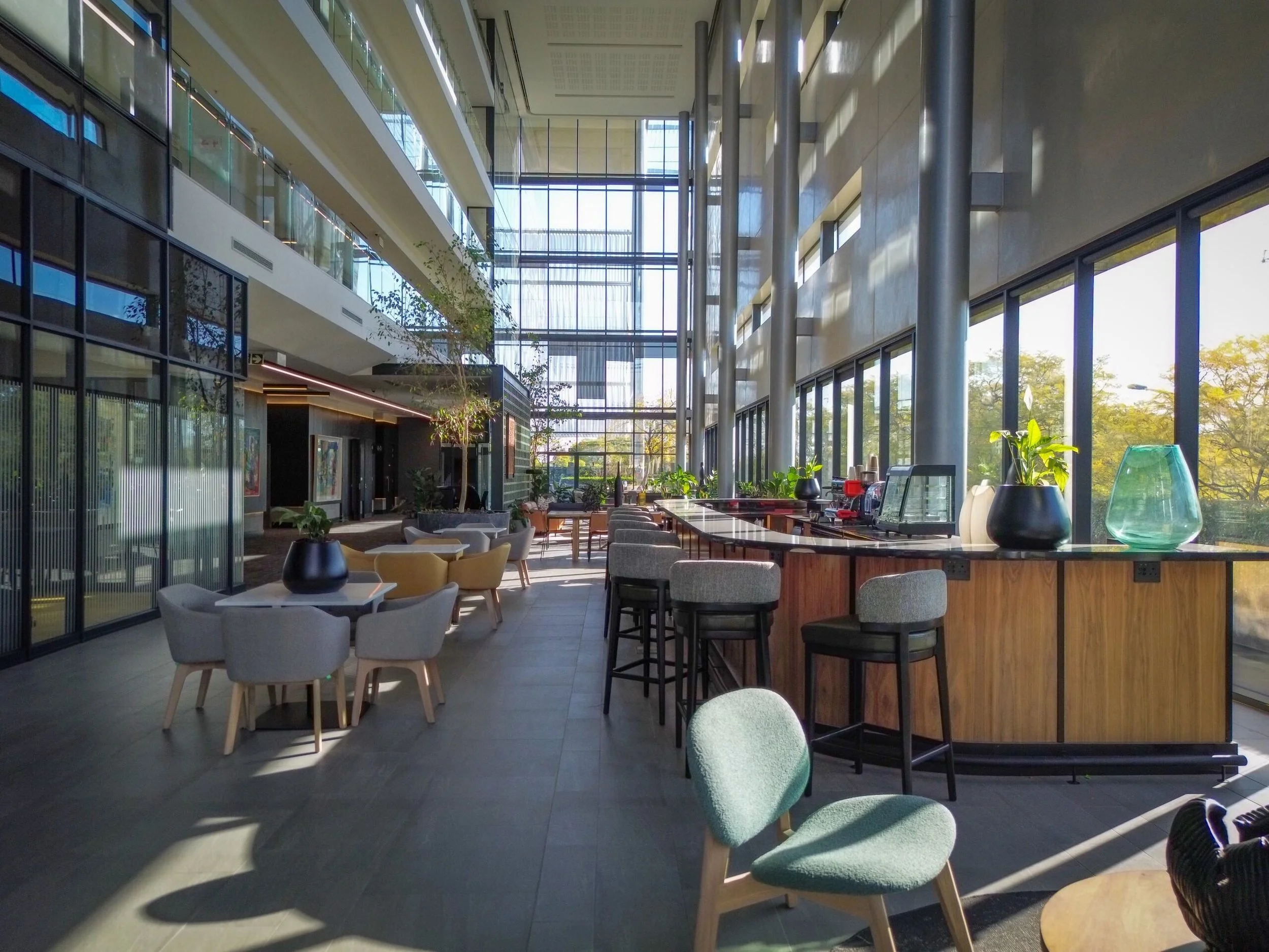 Interior view of a modern cafe or lounge area with large floor-to-ceiling windows, bar seating, and various chairs and tables, decorated with potted plants and artwork.
