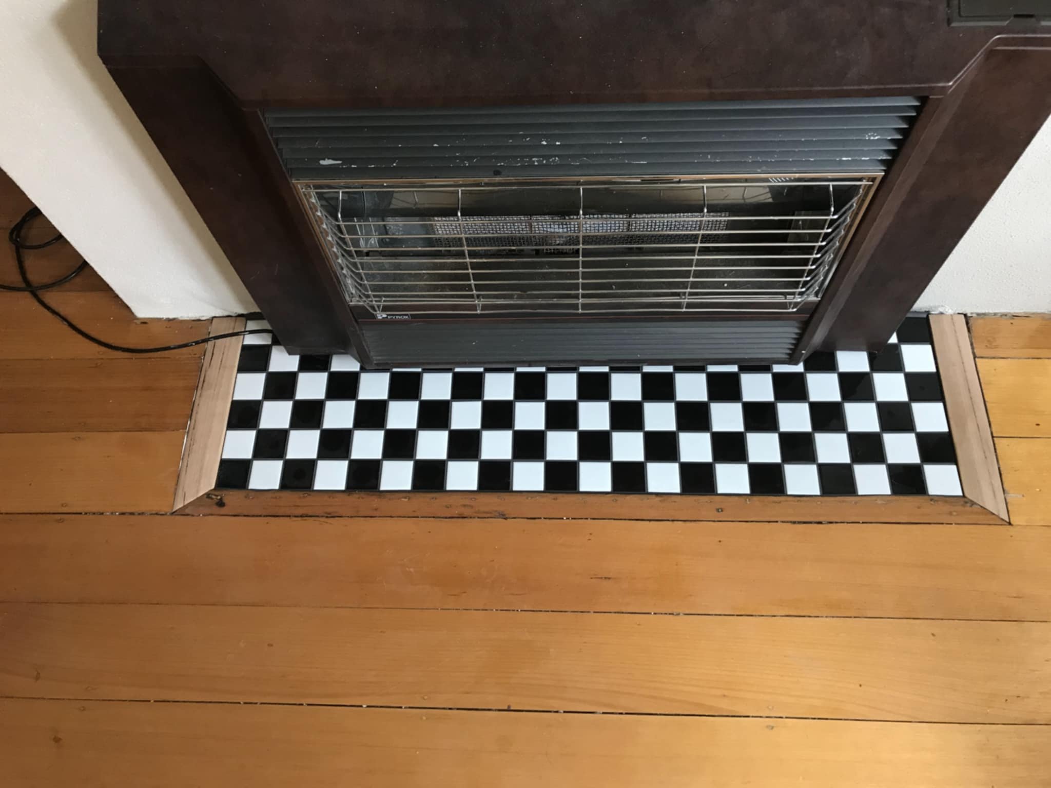 A heater installed on a wooden floor with a black and white checkered tile pattern in front of it.