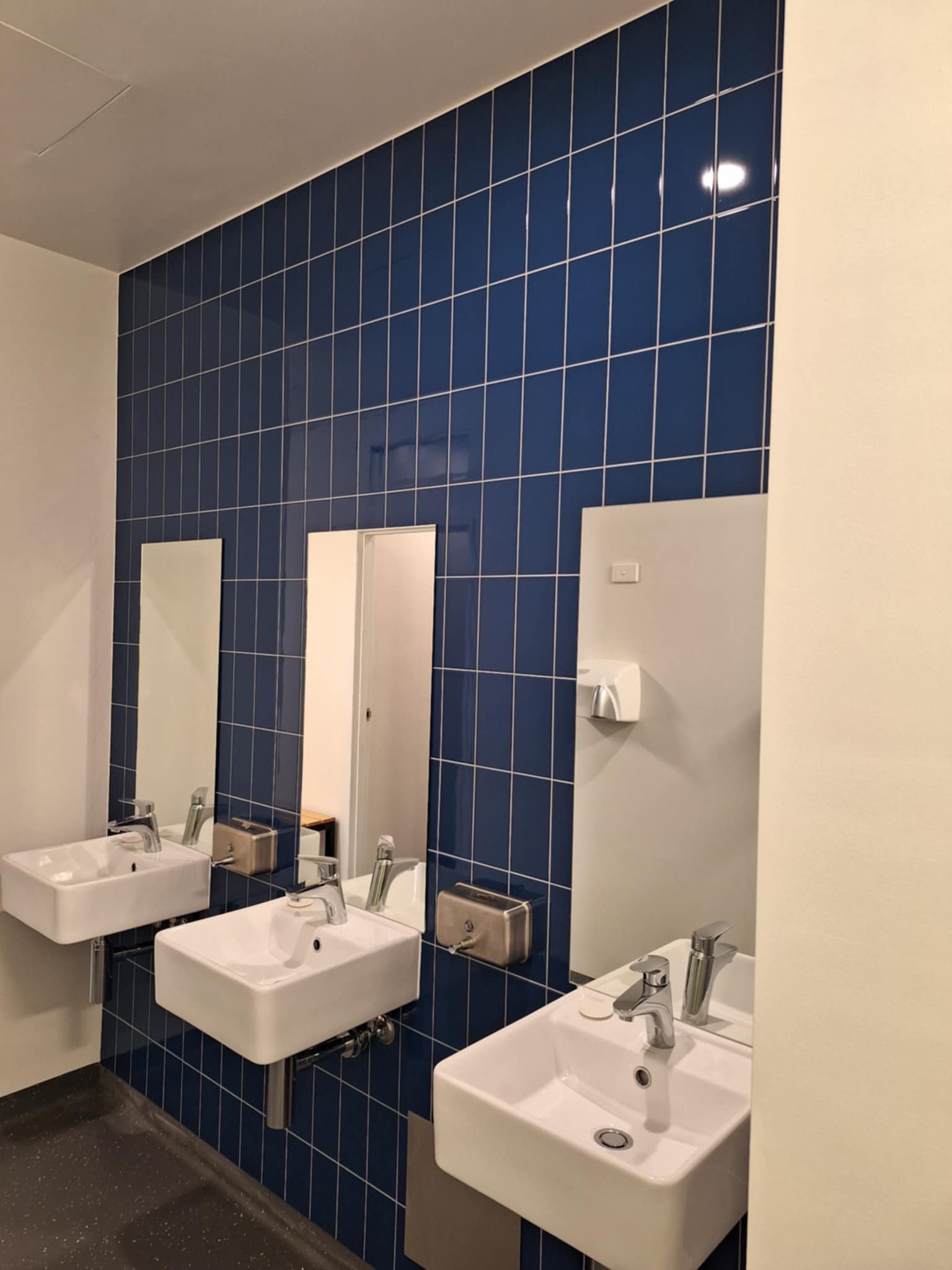 Public restroom with blue tiled wall, two white sinks, mirrors, and metallic soap dispensers.