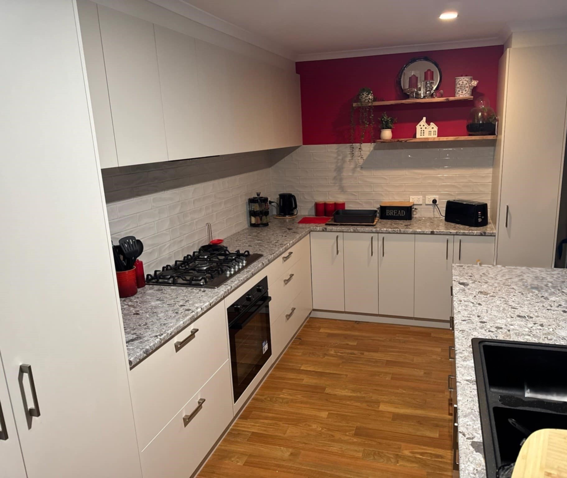 Modern kitchen with gray countertops, white cabinets, and red accents. Features include a gas stove, oven, coffee maker, toaster, and decorative items on shelves.