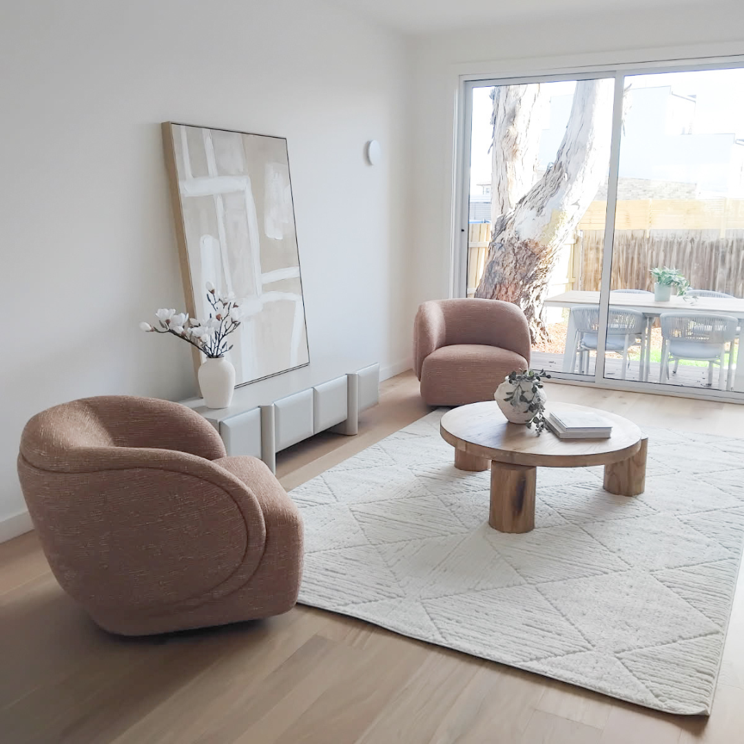 Minimalist living room with two pink armchairs, a round wooden coffee table, a white rug, a large abstract painting, a vase with white flowers, and sliding glass doors leading to an outdoor patio with a tree and garden furniture.