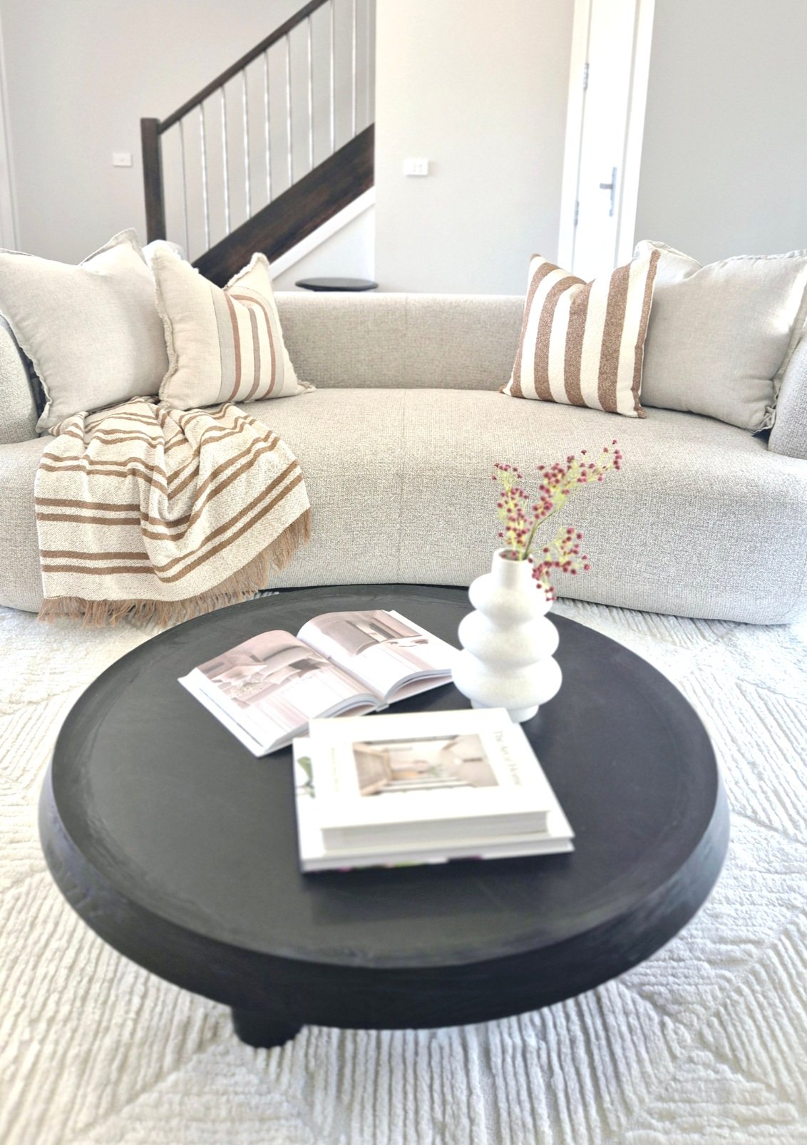 Modern living room with a gray sofa, striped pillows, and a striped throw blanket. A black round coffee table holds a white decorative vase with sprigs and two open books. Carpeted floor and staircase in the background.