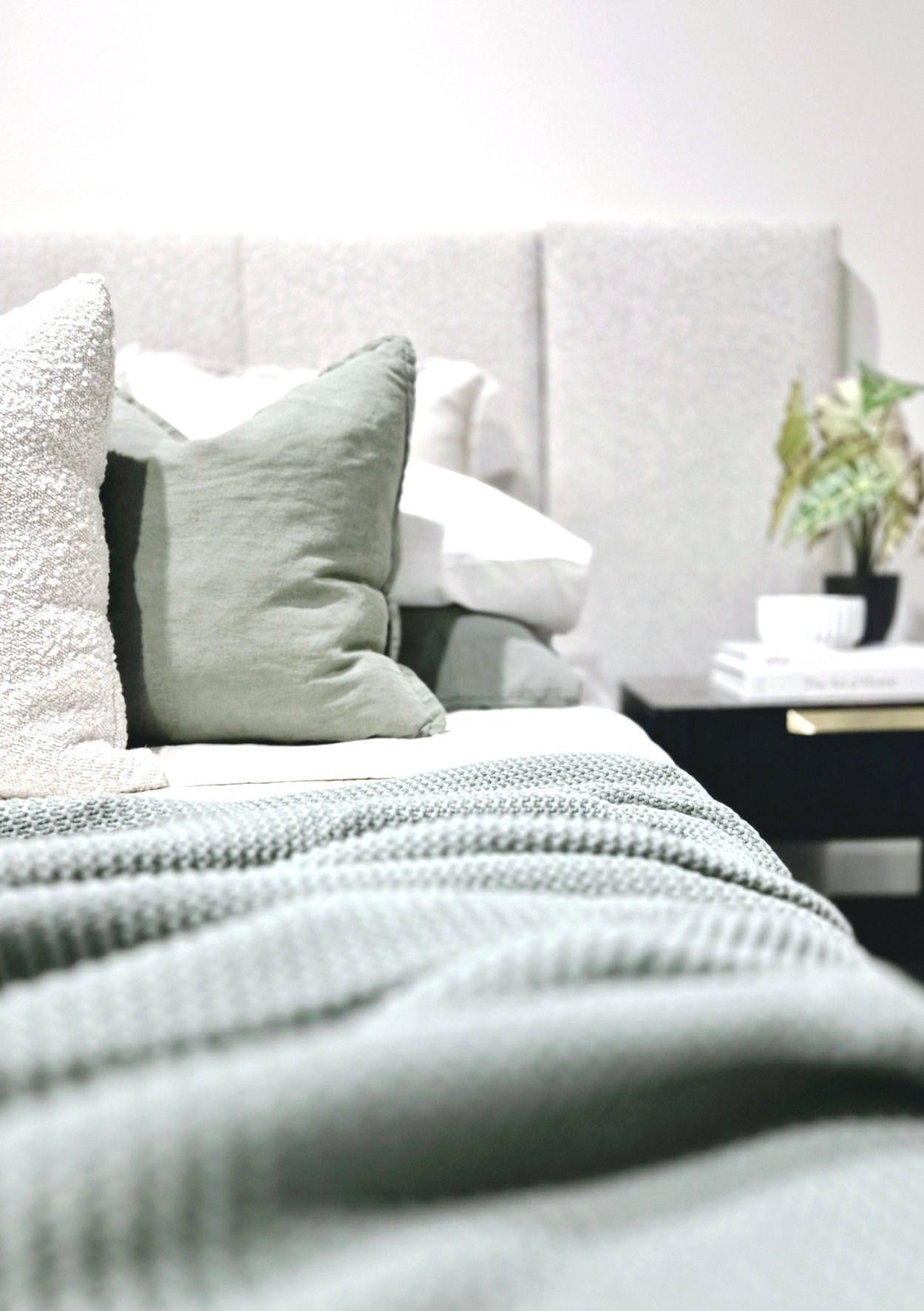 Close-up of a neatly made bed with textured blankets and pillows, against a soft upholstered headboard. There is a bedside table with books and a potted plant.