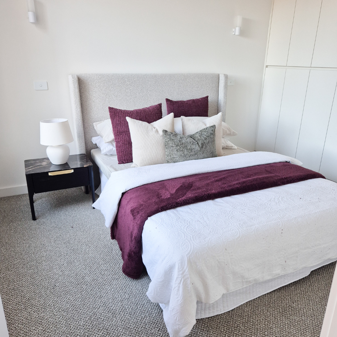 A neatly made bed with white bedding, a maroon throw blanket, and multiple pillows in shades of maroon, white, and gray. A black bedside table with a white lamp is on the left, and a white wall with built-in closet cabinets is in the background.