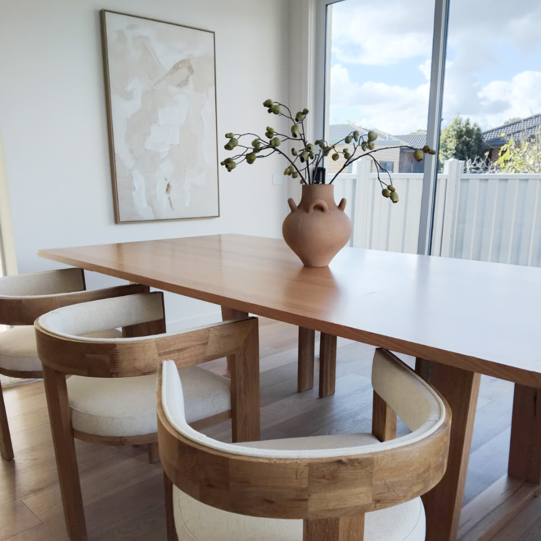 A wooden dining table with four upholstered chairs in a bright room, a large abstract painting on the wall, and a vase of branches on the table next to a sliding glass door showing a cloudy sky and house rooftops outside.