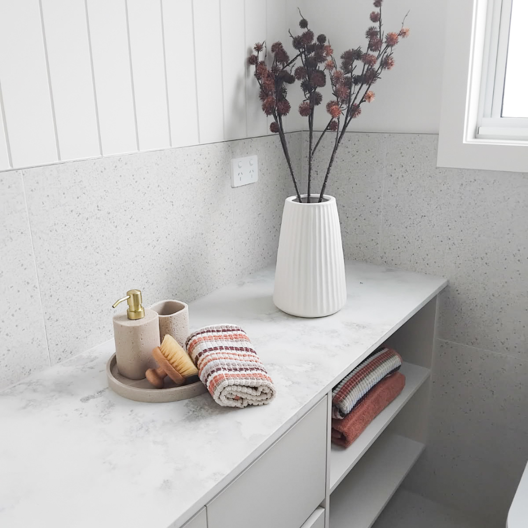 A white marble countertop with a beige soap dispenser, a toothbrush holder, a brush, and a rolled towel. A tall white vase with dried flowers, and some folded towels on a lower shelf. There is a window on the right side of the image.