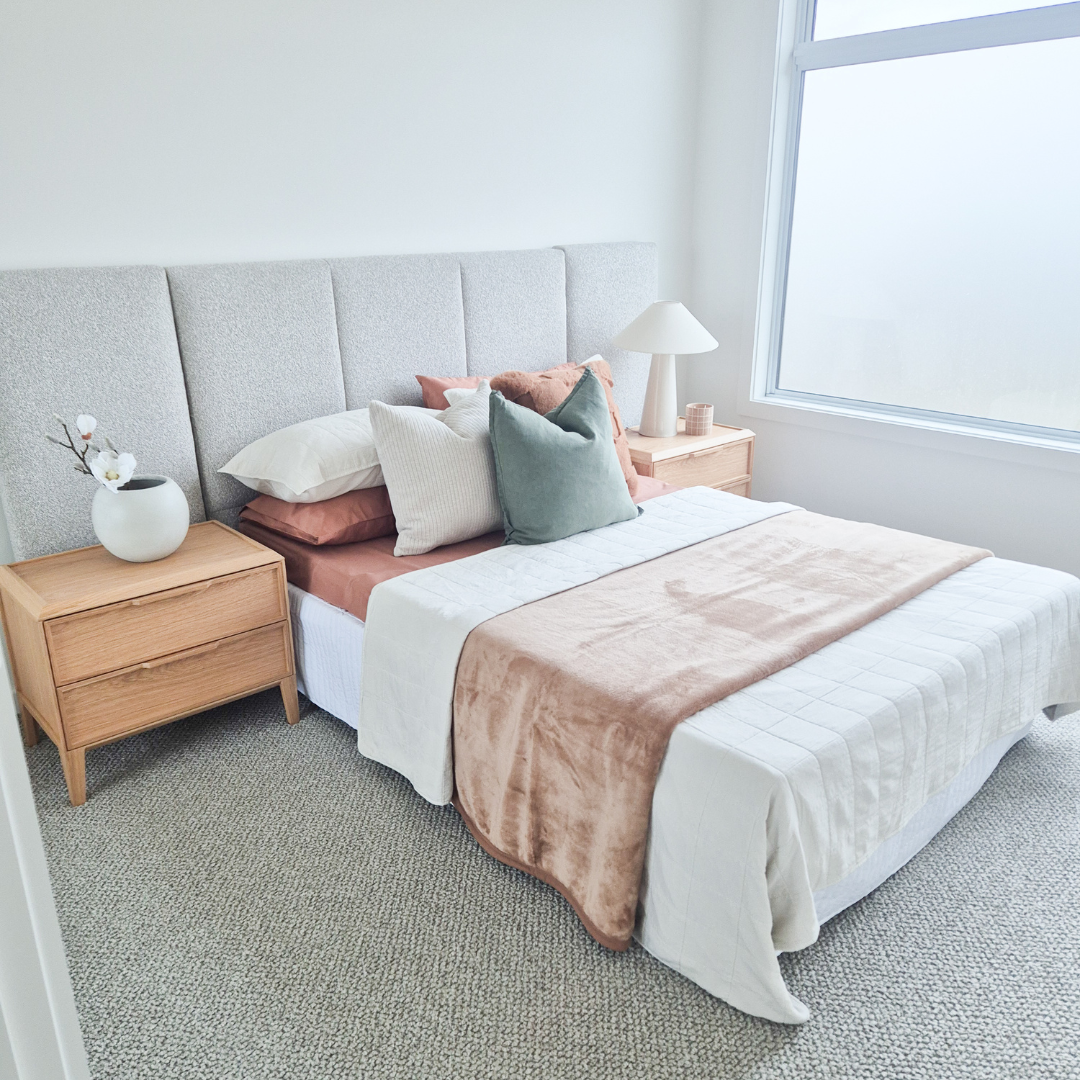 A neatly made bed with white, pink, and green pillows, two light wooden nightstands with lamps, a vase with flowers, a window, and beige carpet in a bright bedroom.