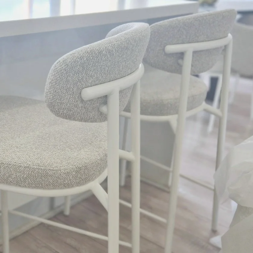 Two modern bar stools with white metal frames and light gray upholstered seats and backs, in a room with wooden flooring.