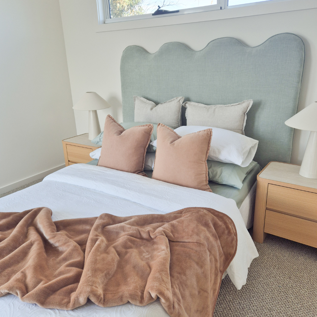 A neatly made bed with a pastel green upholstered headboard, surrounded by beige and pink pillows, and a white blanket with a brown plush throw at the foot of the bed. Two small wooden nightstands with white lamps are on either side of the bed, and a