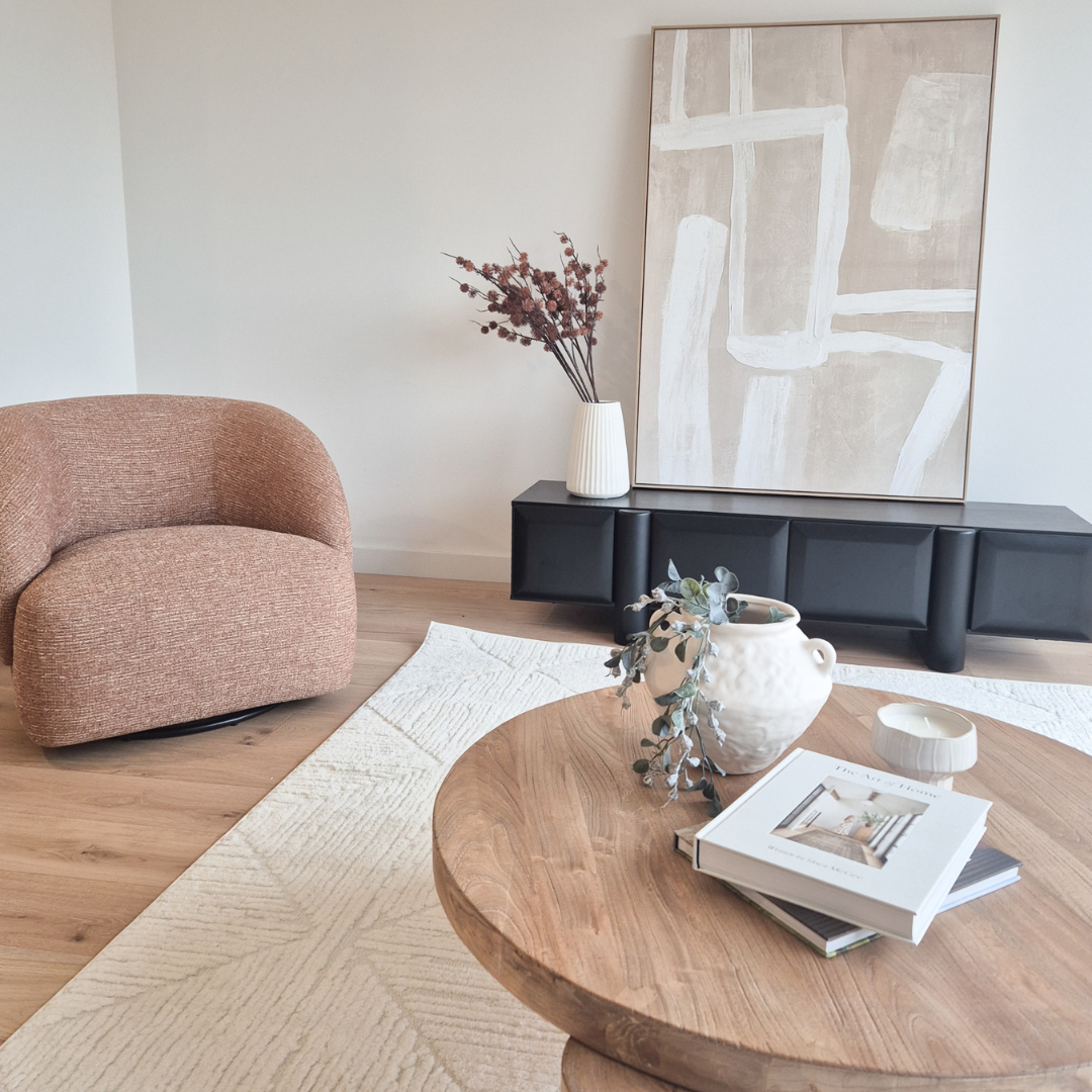 Modern living room with a beige armchair, a black cabinet with a white vase and pink flowers, a round wooden table with books and a white vase with greenery, a neutral abstract painting on the wall, and a light-colored area rug on wood flooring.