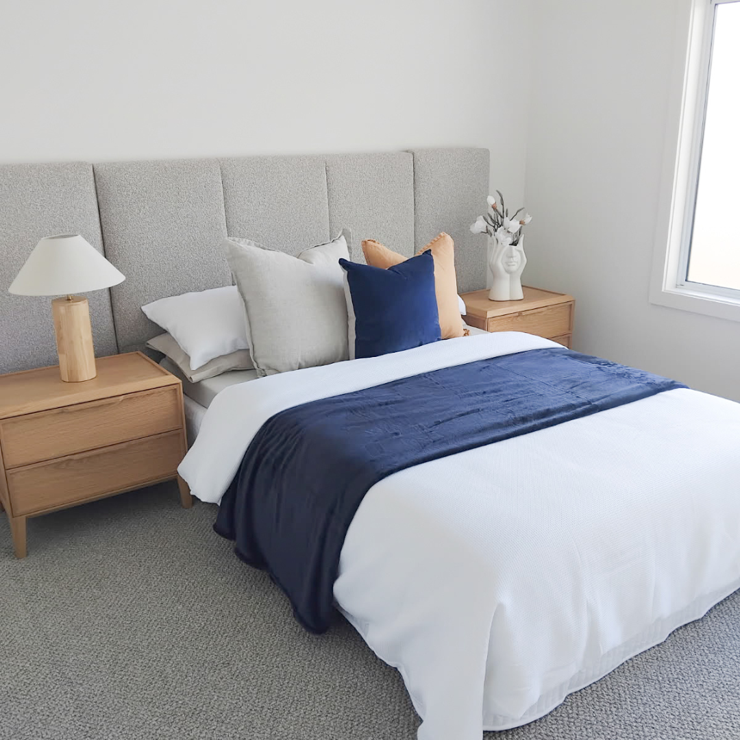 A neatly made bed with a gray upholstered headboard, white bedding, a navy blue throw blanket, and decorative pillows in white, navy, and tan. Two wooden nightstands with a white lamp and a white vase with flowers sit on either side of the bed. A win
