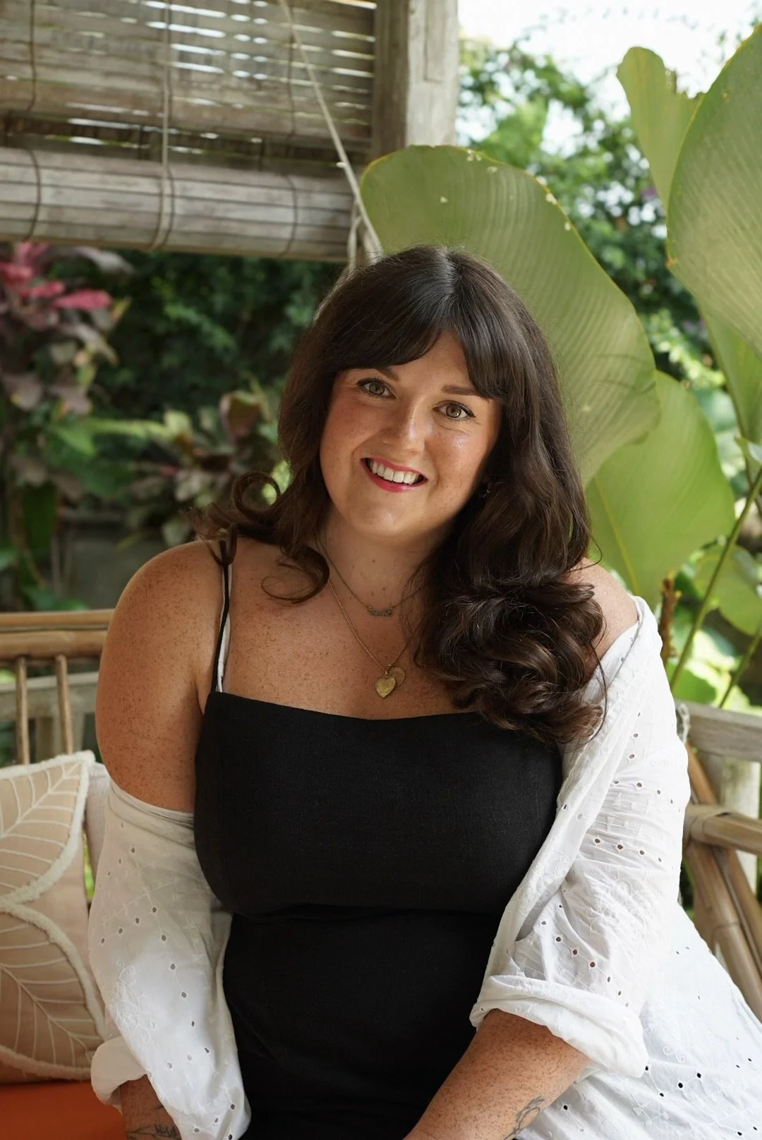 Woman with long brown hair sitting on a patio, wearing a black dress and a white cover-up, surrounded by greenery and decorative plants.
