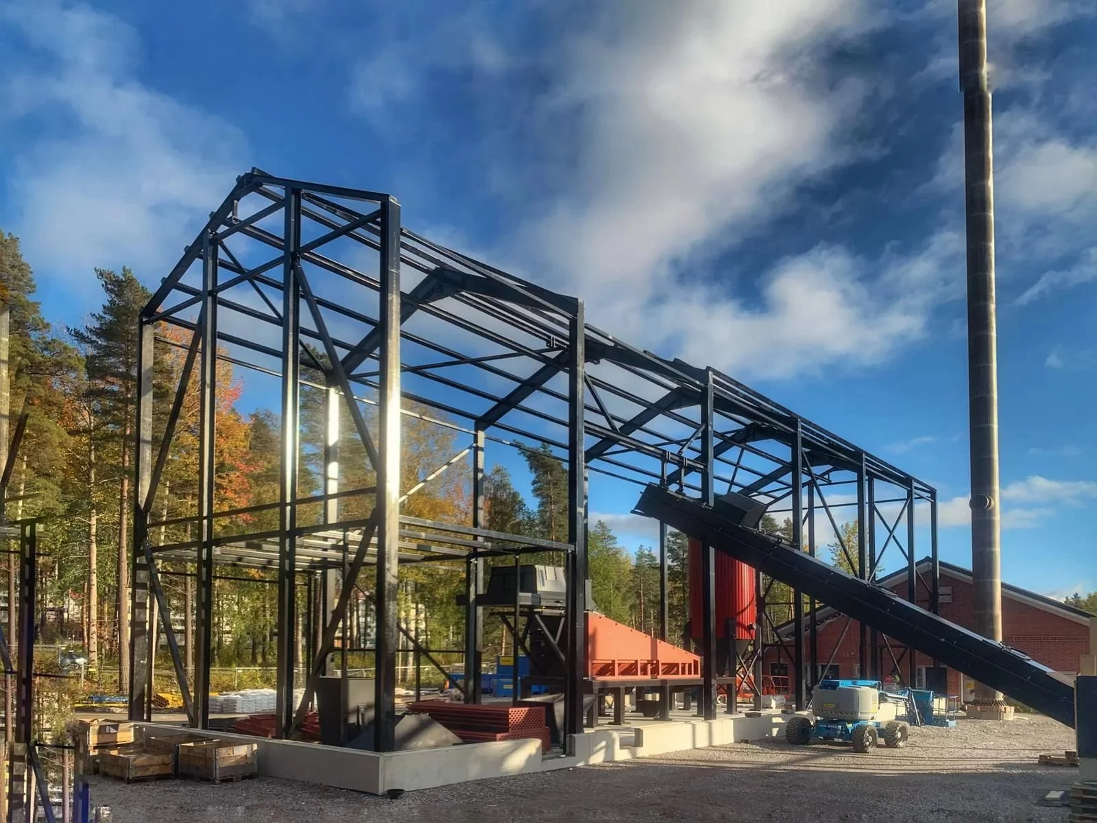 Building under construction with a metal framework, a red hopper, conveyor system, construction equipment, and a partly cloudy sky.