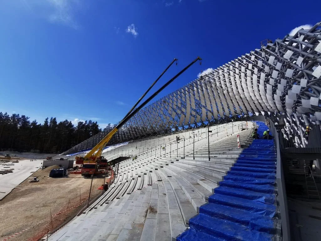 Under construction grandstand with metal framework and seats, and cranes working on the structure under a blue sky.