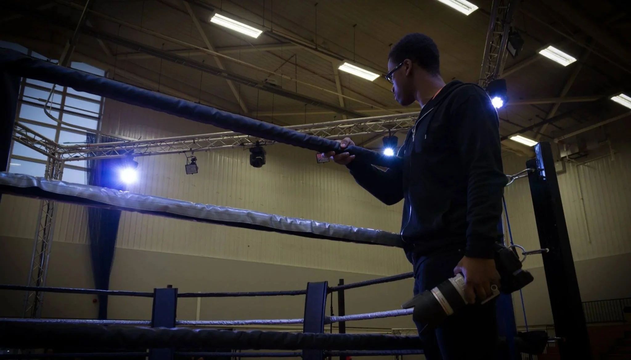 A man standing in a boxing ring in an indoor gym, holding a camera and looking at a phone, with bright lights overhead.