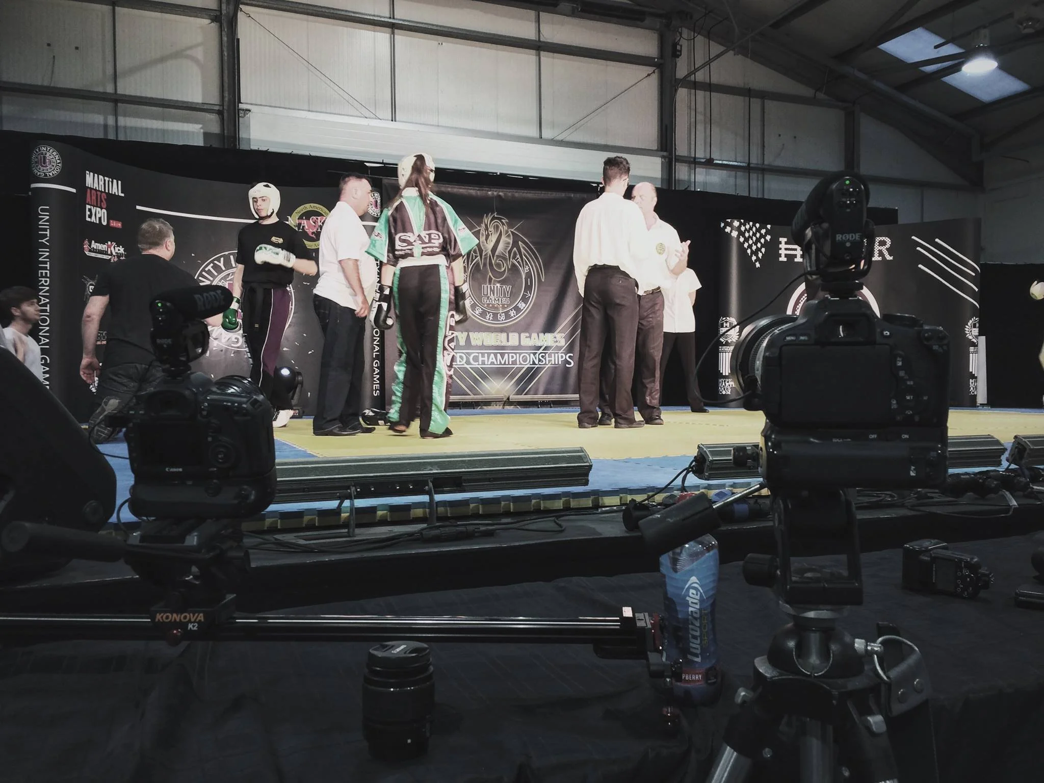A martial arts event with participants and officials on a stage, with cameras and filming equipment set up in front.