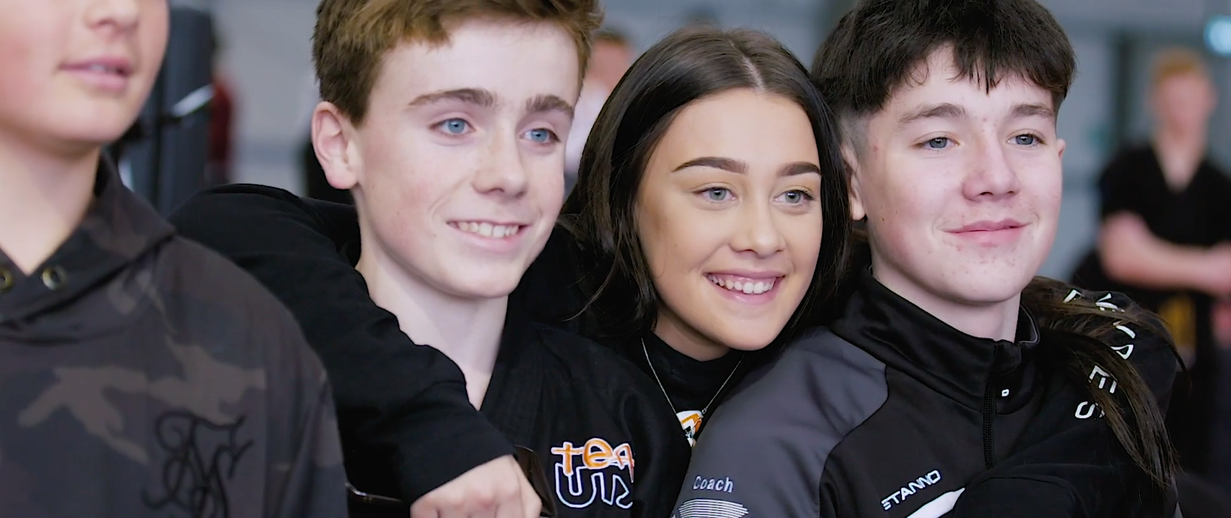 Four teenagers smiling and posing together indoors, wearing black and gray sports jackets, with their arms around each other.