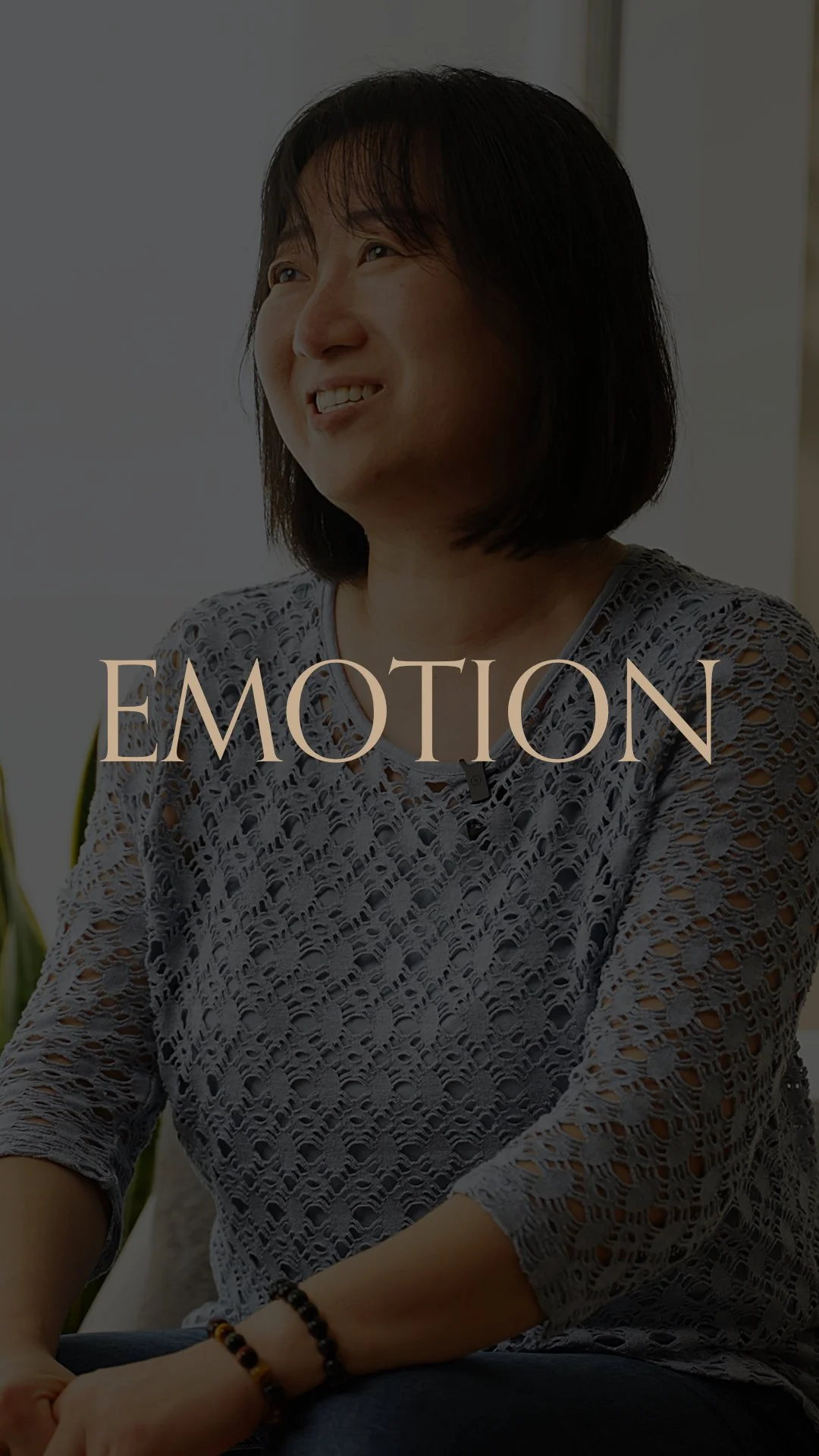 A woman with dark hair smiling and gazing to her left, wearing a lacy grey top, with the word 'EMOTION' overlayed on the image.