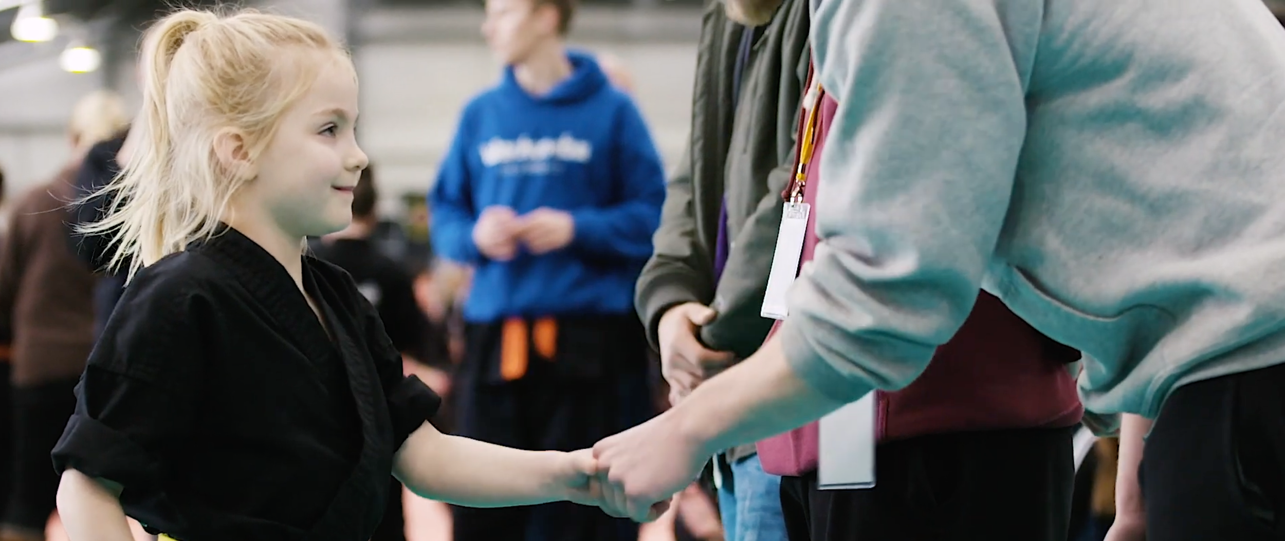 A young girl with blonde hair tied back, wearing a black martial arts uniform, shaking hands with an adult wearing a gray hoodie. Several people are in the background of the indoor setting.