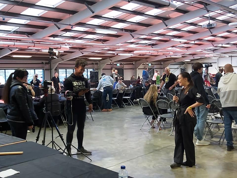 People gathering and working with cameras and microphones in a large indoor space, possibly a sports martial arts tournament.