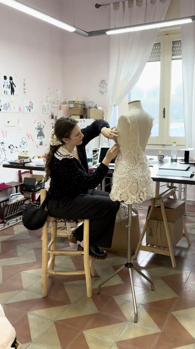 A woman working on a lace dress on a dress form in a room with a window, art on the wall, and sewing supplies on tables.
