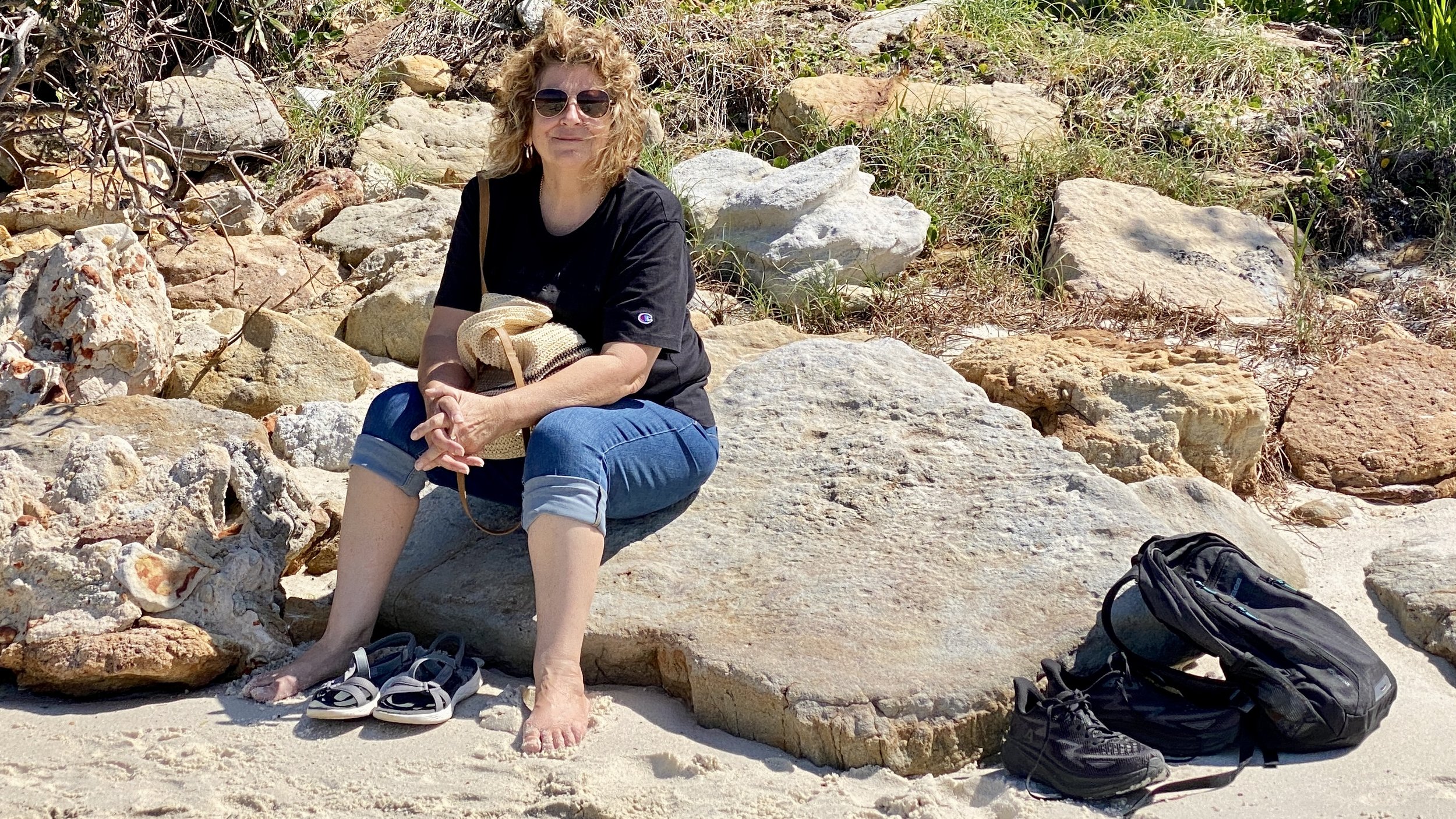 Woman sitting on a large rock on a sandy beach, wearing sunglasses, black t-shirt, and rolled-up jeans, with a backpack, black sneakers, and sandals nearby, surrounded by rocks and grass.