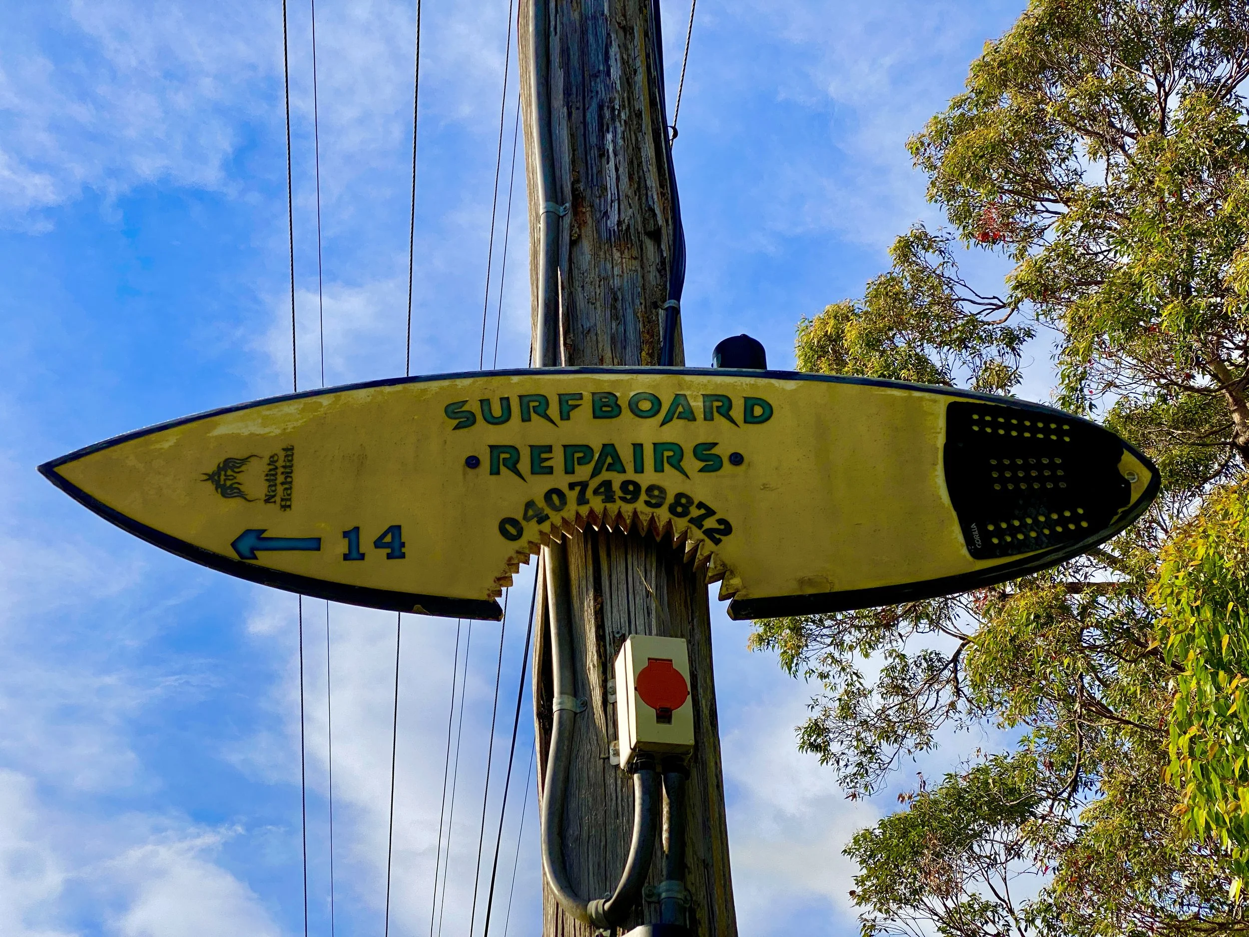 Yellow surfboard-shaped sign on a wooden pole advertising surfboard repairs, with a phone number, an arrow pointing left with the number 14, and a logo of a black seahorse.