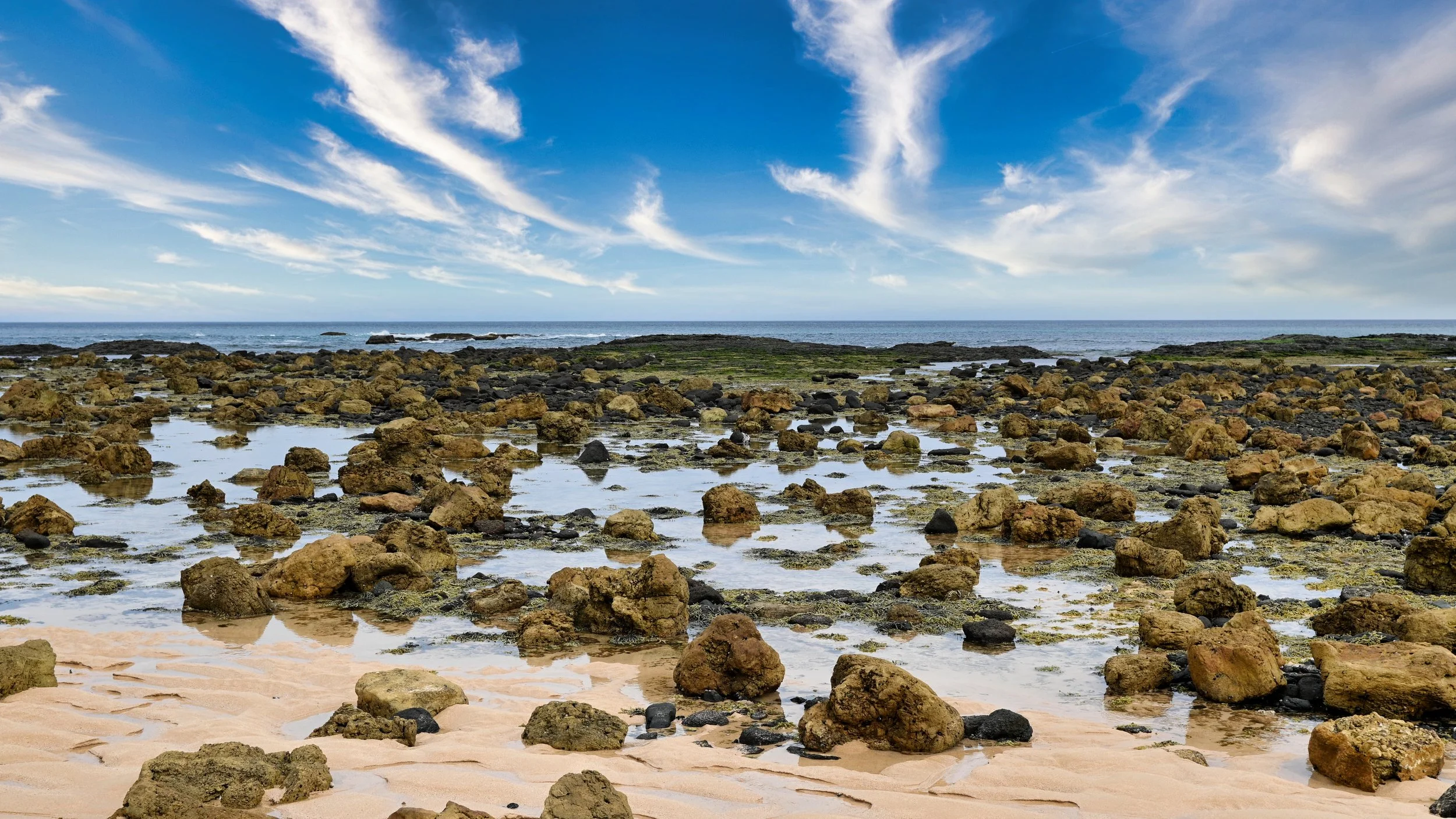 A rocky shoreline with shallow water, sand, the ocean in the distance, and a partly cloudy sky.