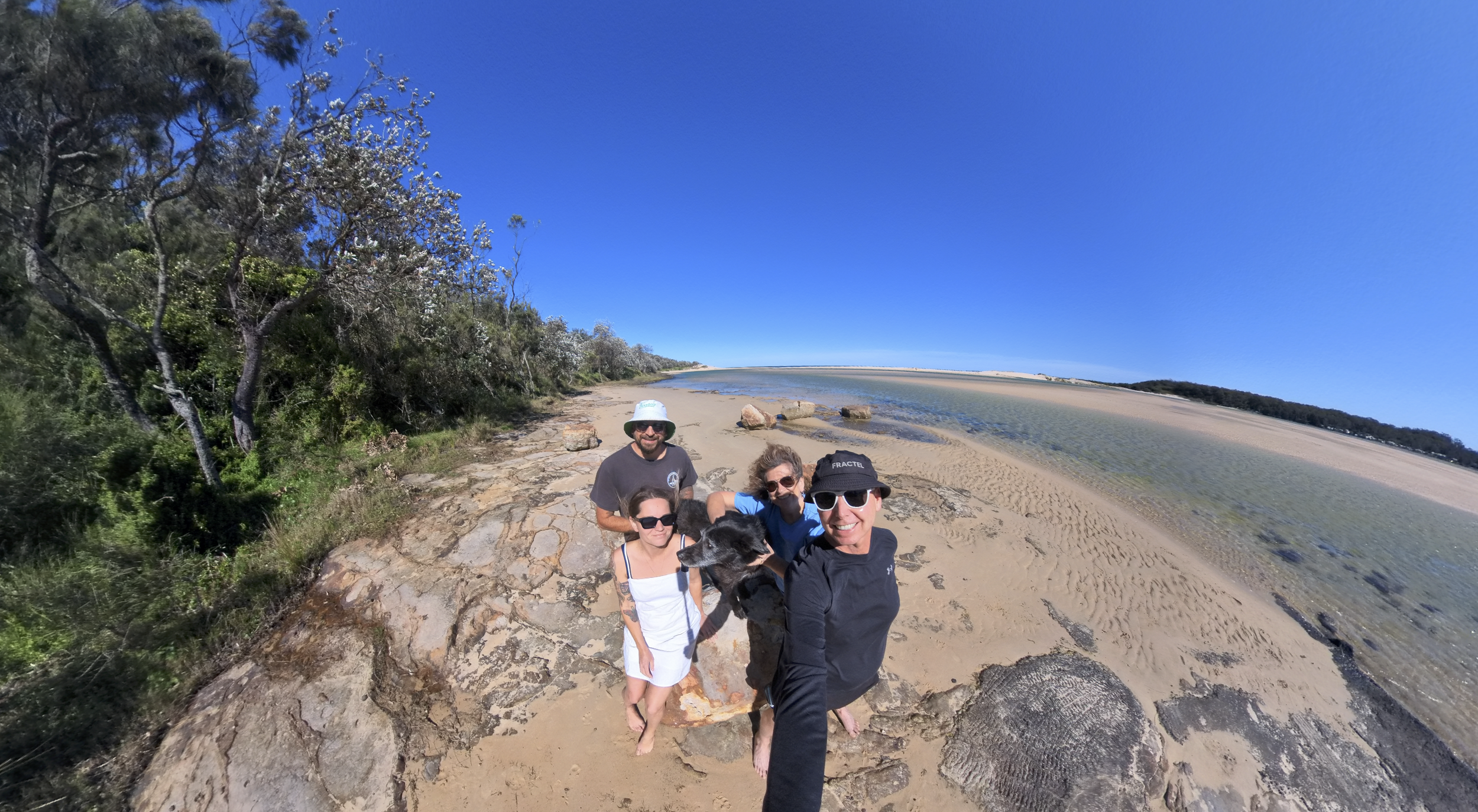 Group of five people and a dog taking a selfie on a rocky and sandy beach under a clear blue sky with trees on the left and water on the right.