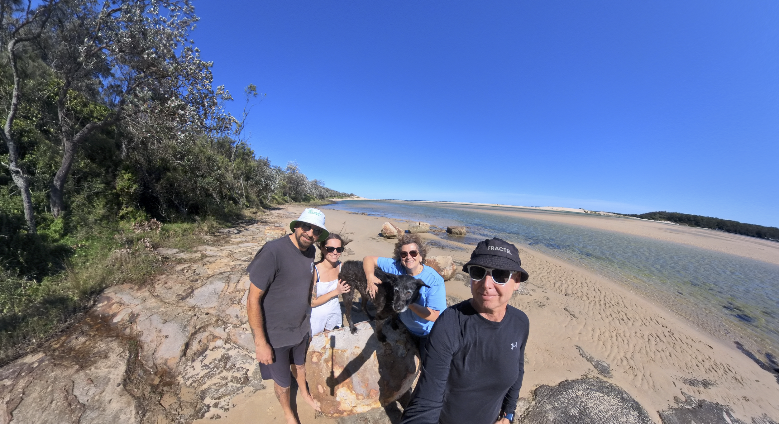 Four people and a black dog on a sandy beach with rocks, trees, and a river or estuary in the background under a clear blue sky.