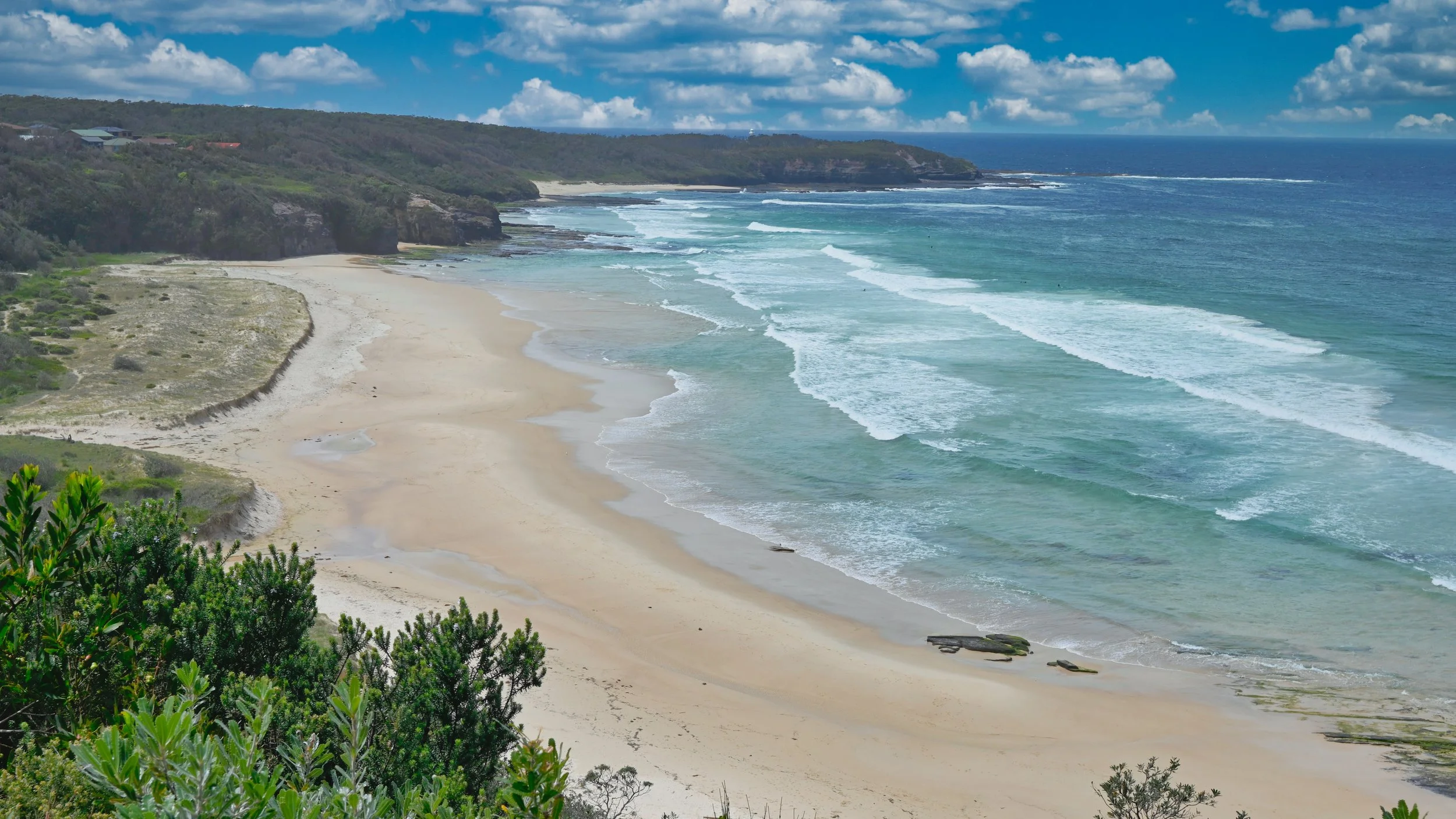 A scenic beach with white sand, ocean waves, rocky cliffs, and green foliage under a partly cloudy sky.