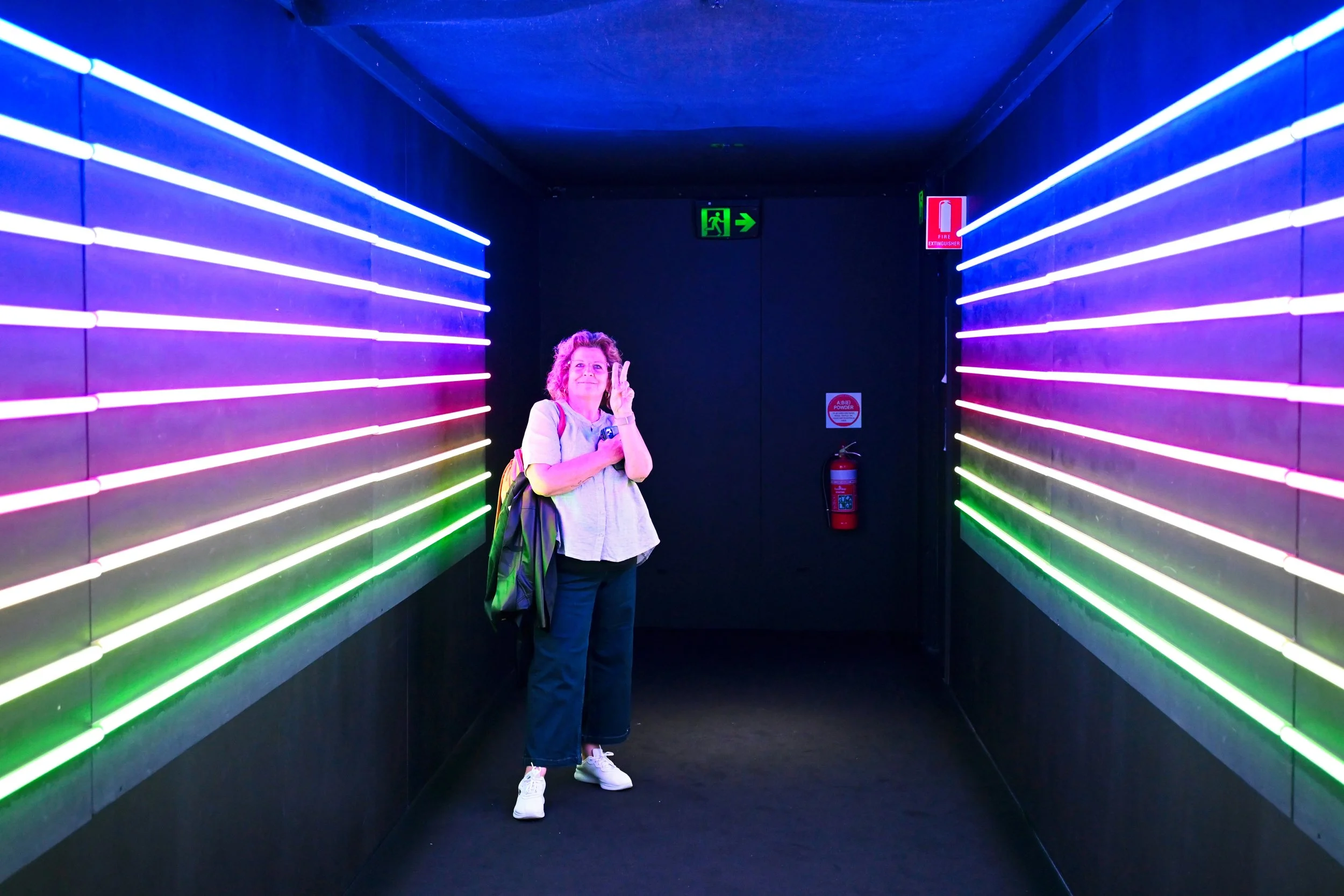 A woman stands in a dark corridor lit with colorful neon lights on the walls, making a peace sign with her right hand.