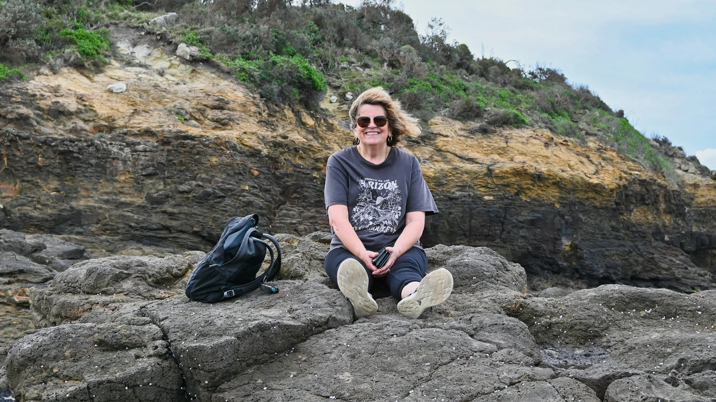 A woman with short curly hair, sunglasses, and a gray T-shirt sitting on a large rock on a coastline, smiling at the camera, with a black backpack beside her.