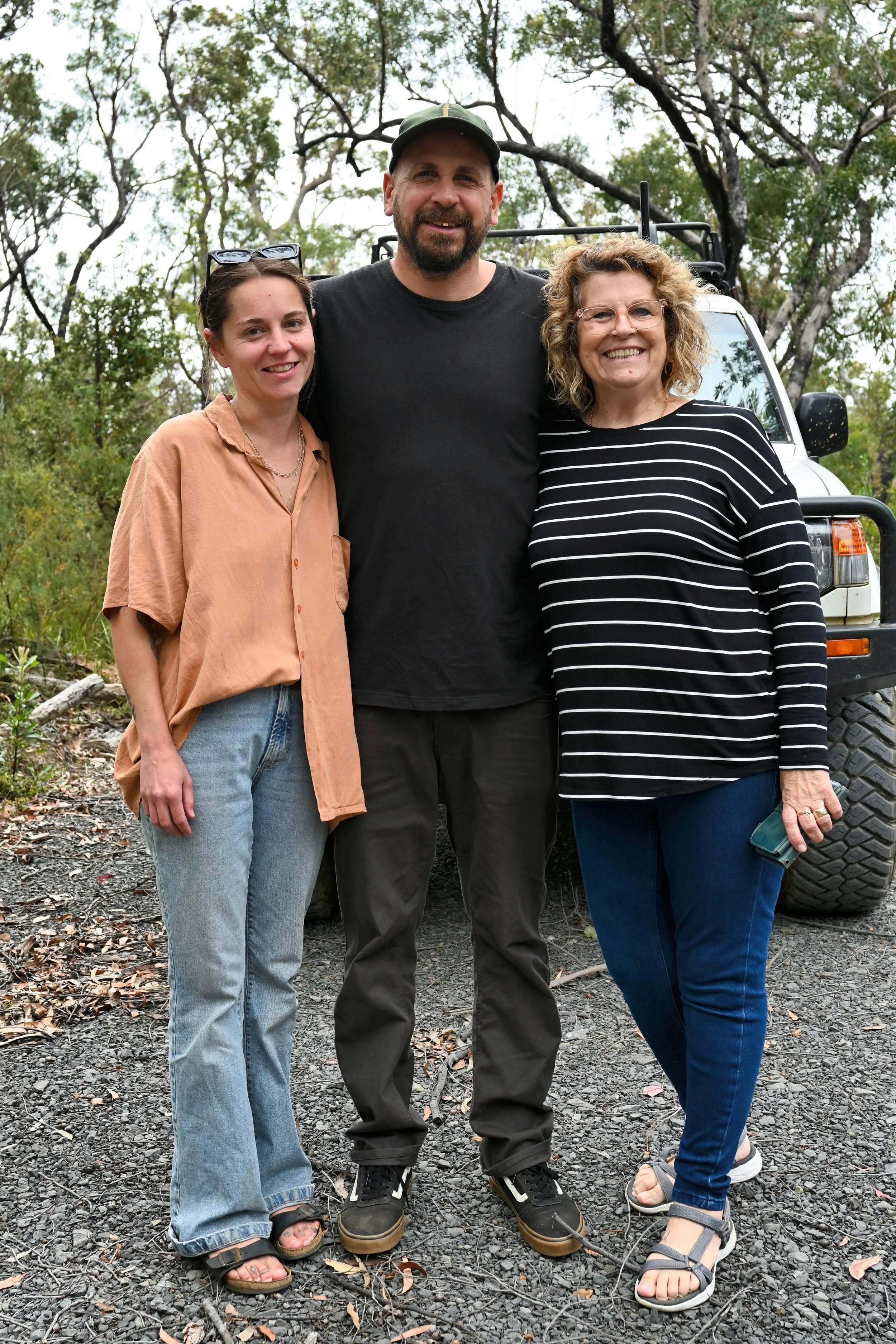Three people standing outdoors in front of a white off-road vehicle surrounded by trees, smiling at the camera. Two women on either side of a man, all dressed casually.