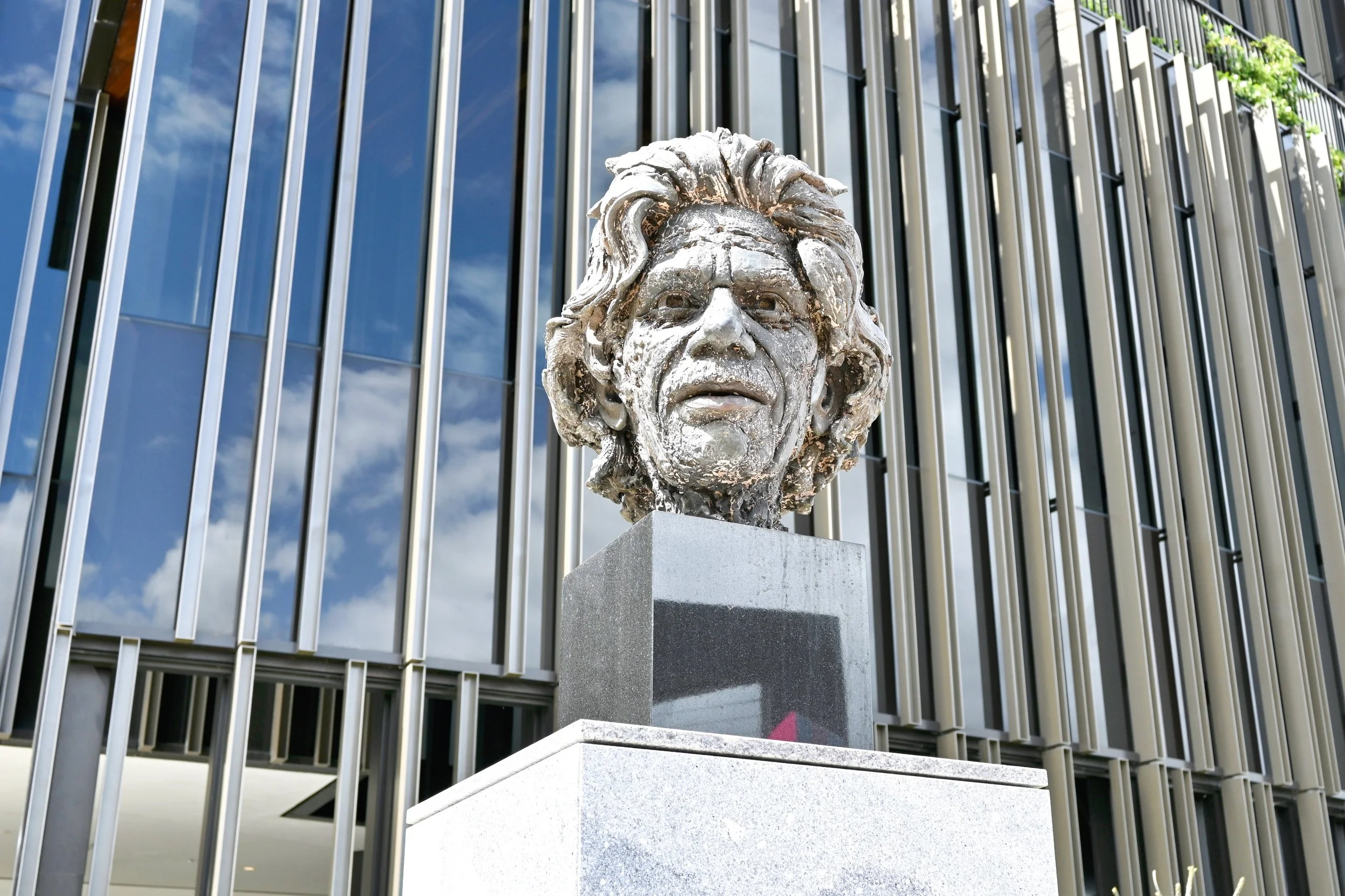Bronze bust of a man with long, wavy hair on a pedestal outside a modern building with glass windows and vertical metal panels.