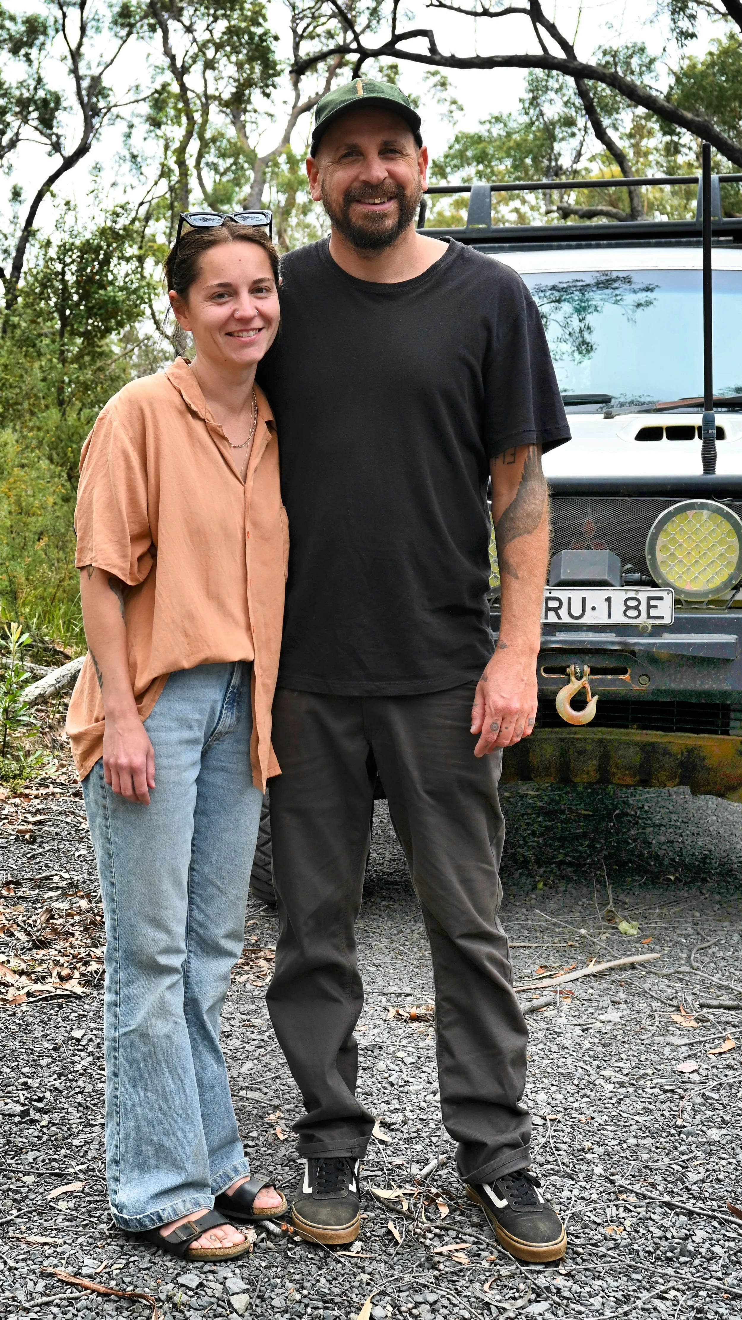 A man and woman smiling outdoors near a vehicle, with trees in the background. The man wears a black t-shirt, dark pants, and a baseball cap. The woman wears a tan loose blouse, light jeans, and sandals. They are standing on a gravel surface.