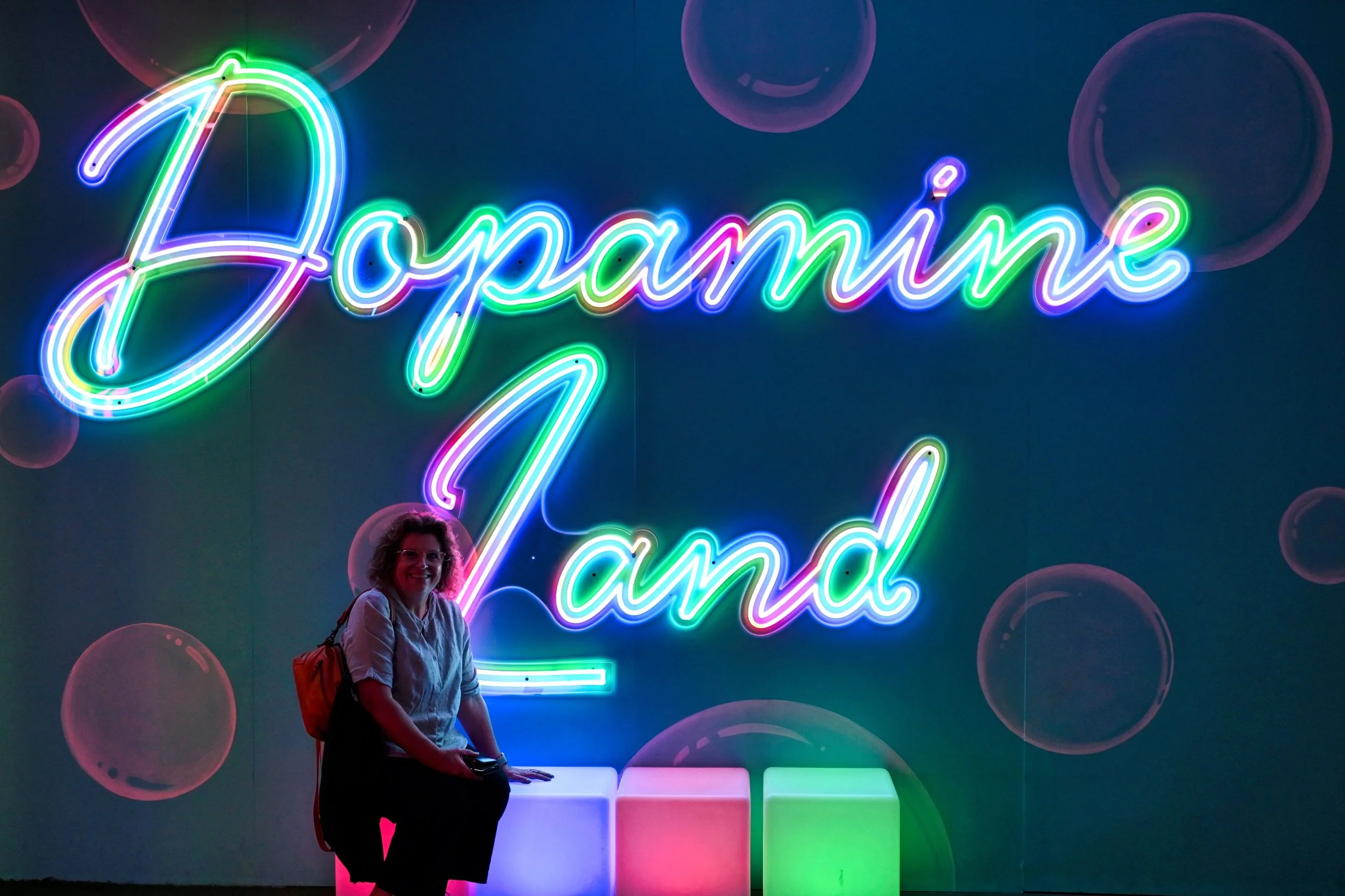 A woman with curly hair, wearing a light-colored shirt and black pants, stands in front of a neon sign that reads 'Joyamine Land' in colorful cursive letters, illuminated against a dark background with floating bubble shapes.