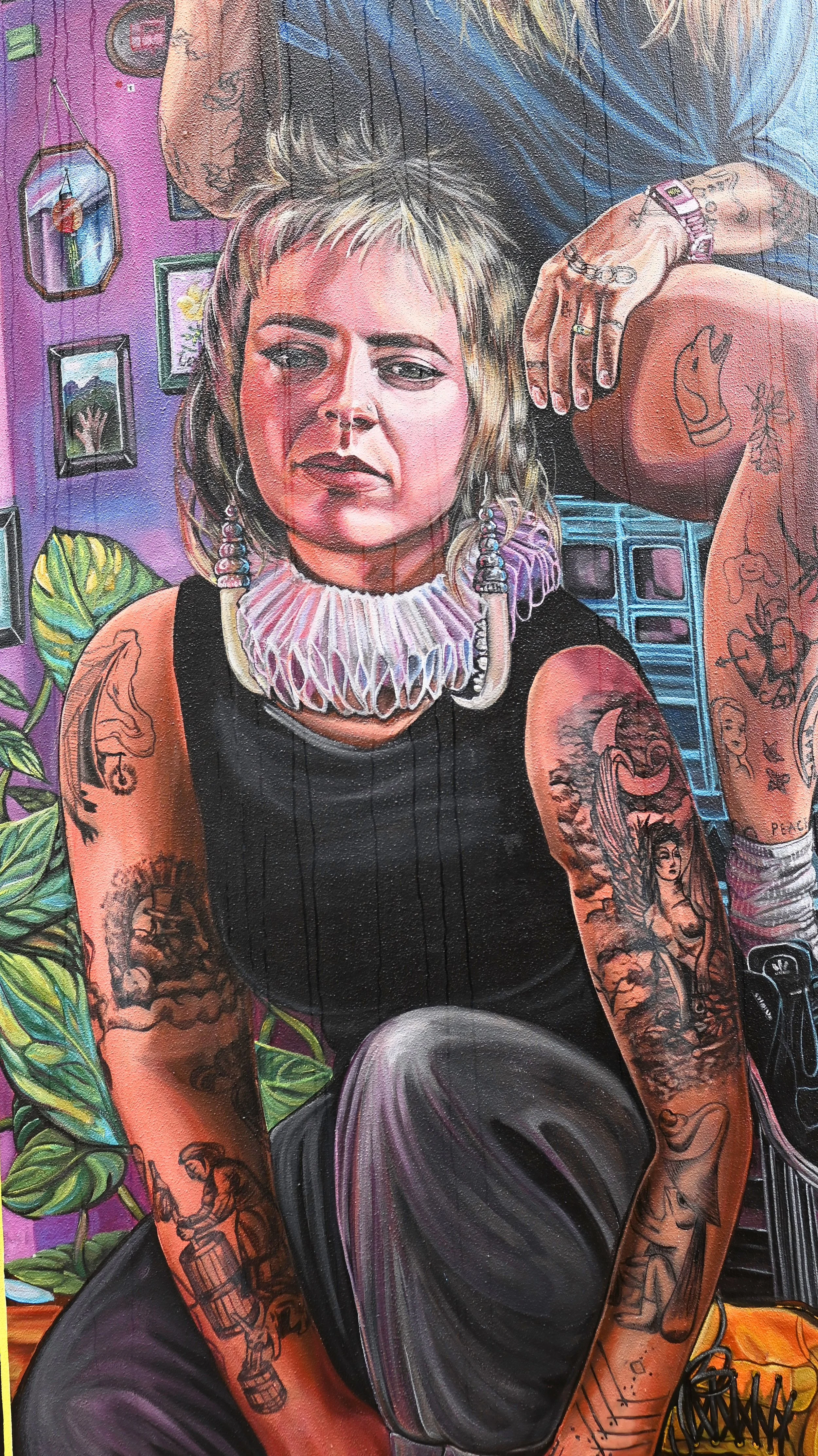 A woman with short, blond hair, tattoos on her arms, and large earrings, sitting in a colorful room with purple walls, various framed pictures, and green plants.