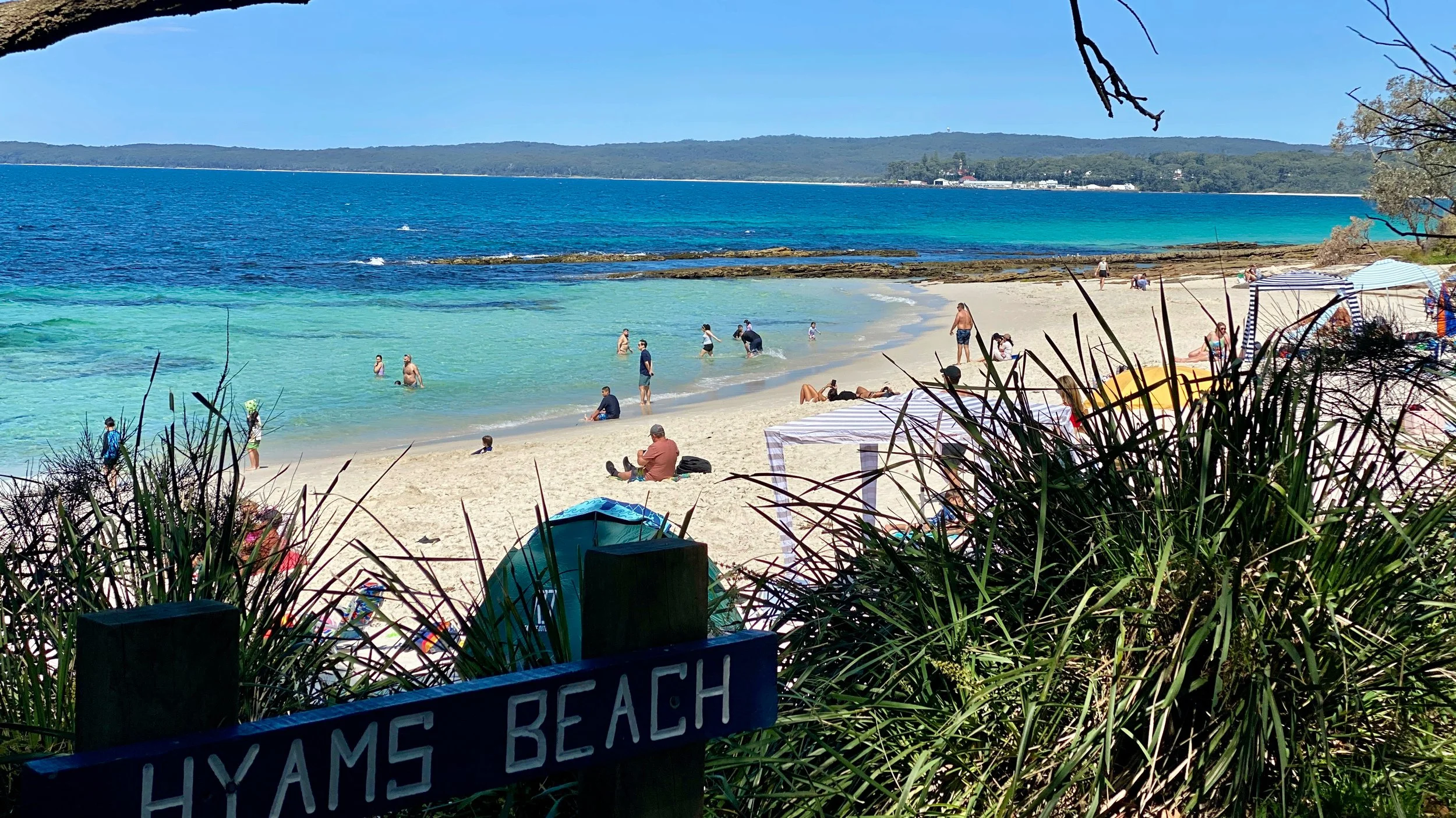Beach scene with people swimming, relaxing, and walking along the sandy shore, with clear blue water, a sign reading 'Hyams Beach' in the foreground, greenery in the foreground, and a distant tree-lined coastline under a blue sky.