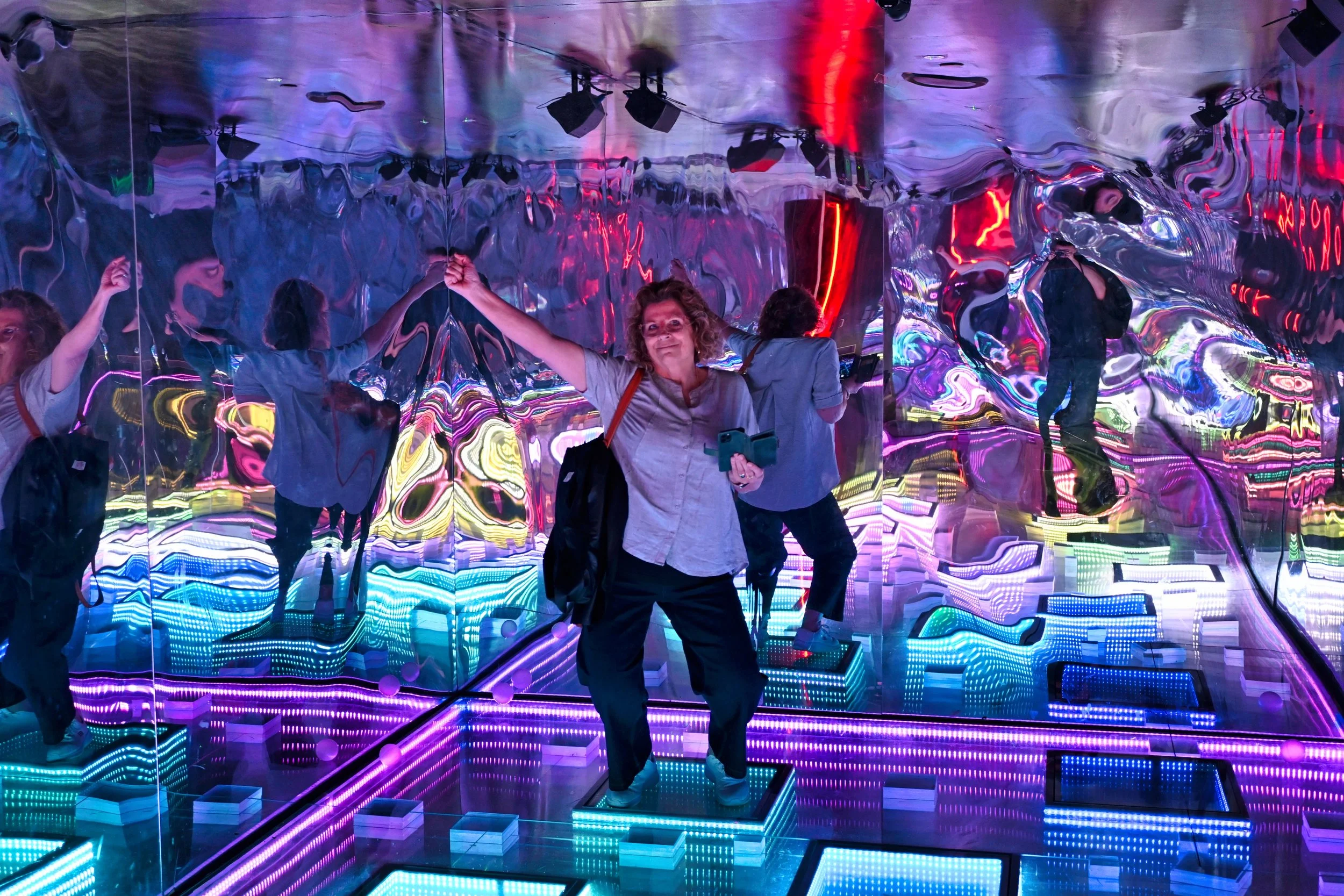 A woman standing on a lit-up platform inside a mirrored, neon-lit room, holding a phone and kicking her right leg up, with other people in the background dancing and taking photos.