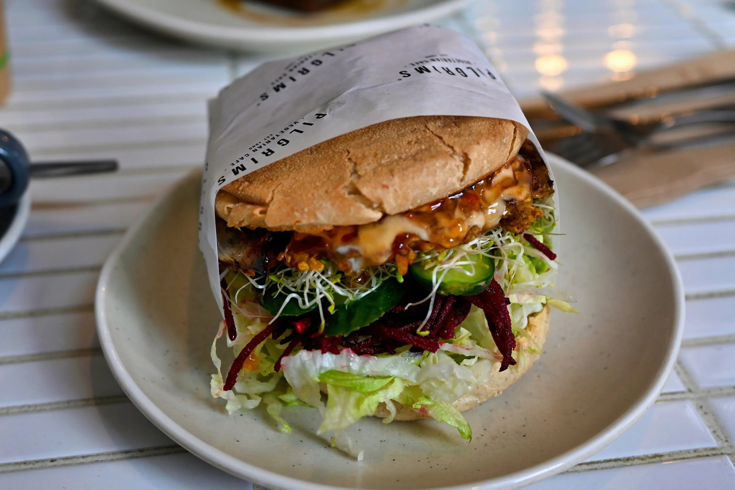 A sandwich wrapped partially in white paper with black text, served on a beige plate. The sandwich contains lettuce, cucumber, shredded beets, a fried patty, melted cheese, and sauce, all inside a crusty bread roll.