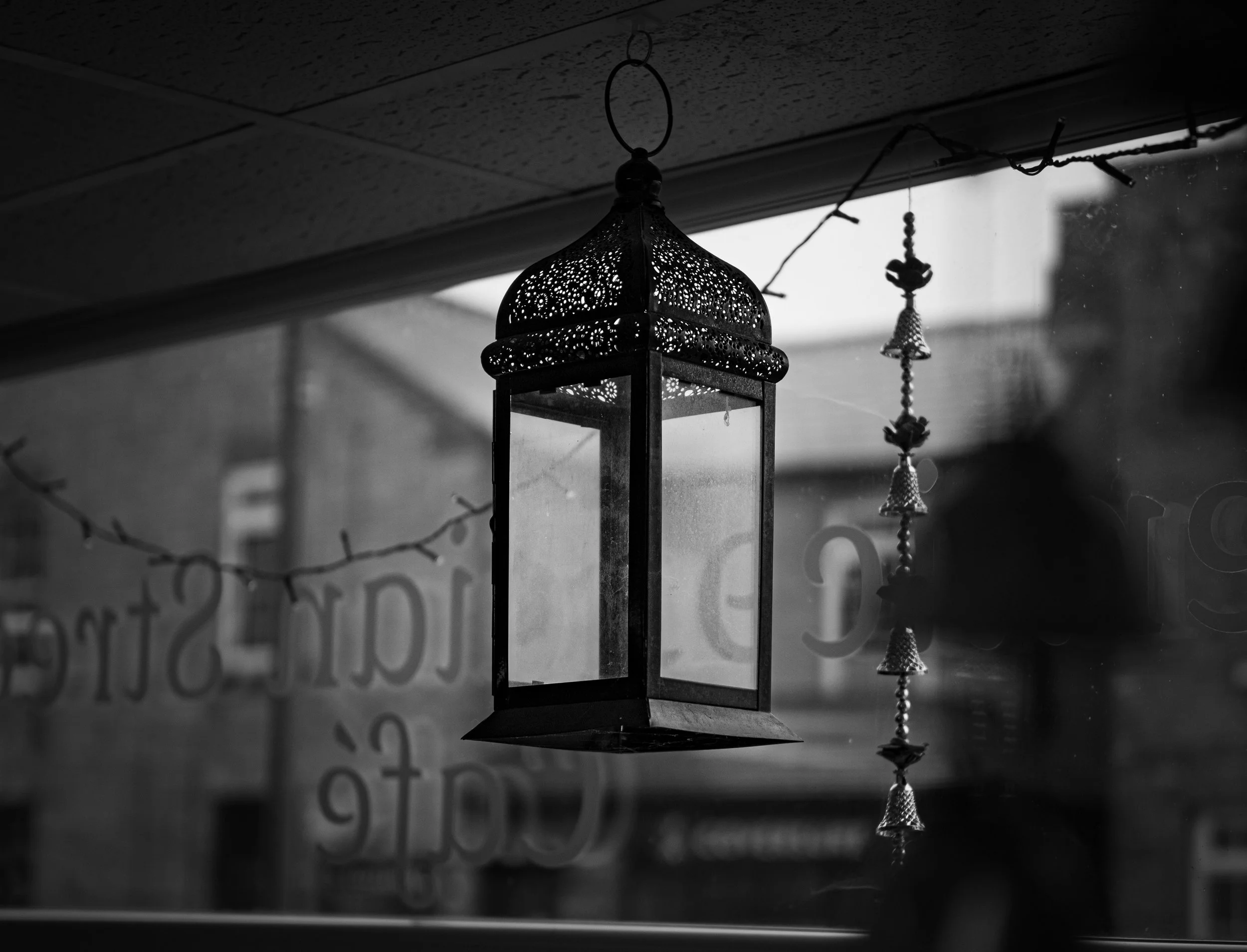 Black and white photo of a decorative hanging lantern with intricate patterns, suspended by a chain, with a string of small decorative bells hanging beside it. The background shows a blurred storefront or window with some text.