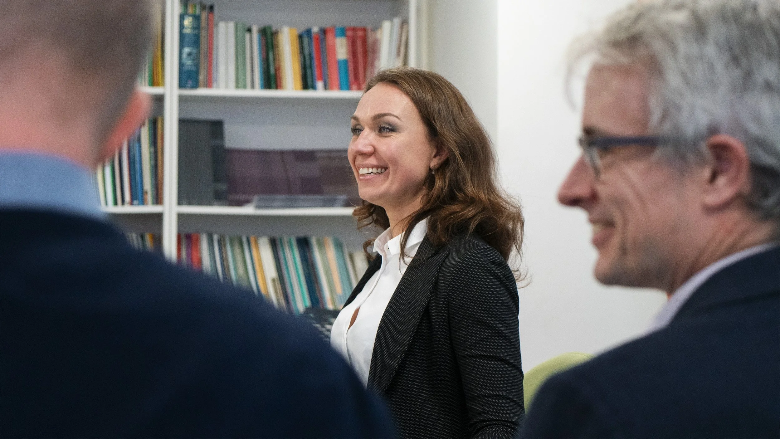Three people in a professional setting, with white bookshelves filled with colorful books in the background. The woman at the center has brown hair and is smiling, wearing a white shirt and black blazer. The man on the right has gray hair, glasses, a