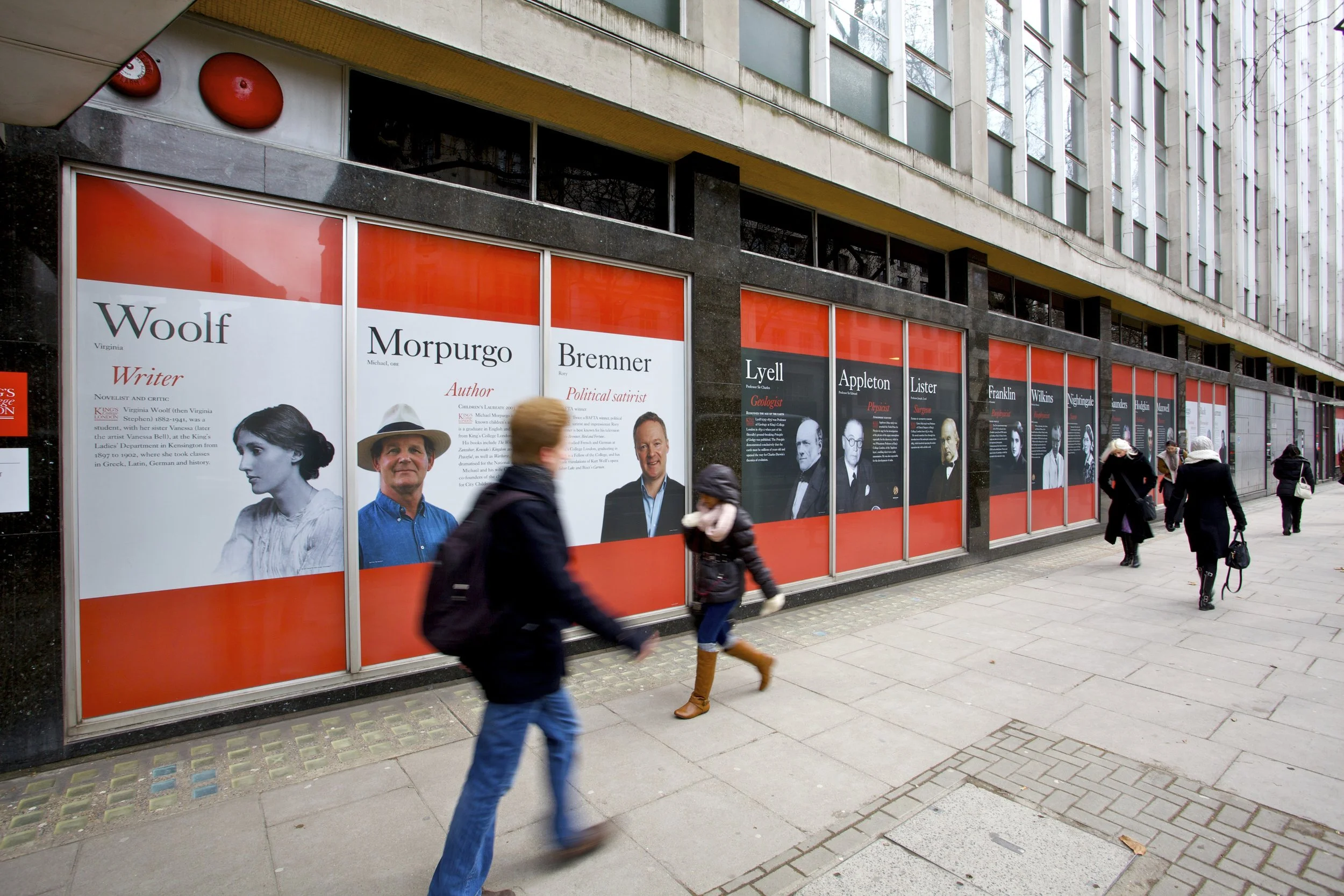 window display with large red and white posters featuring images and bios of various writers and authors, including Woolf, Morpurgo, Bremer, Lyell, Appleton, Lister, Franklin, Wilkins, and Volta. Pedestrians walking on the sidewalk in front of the di